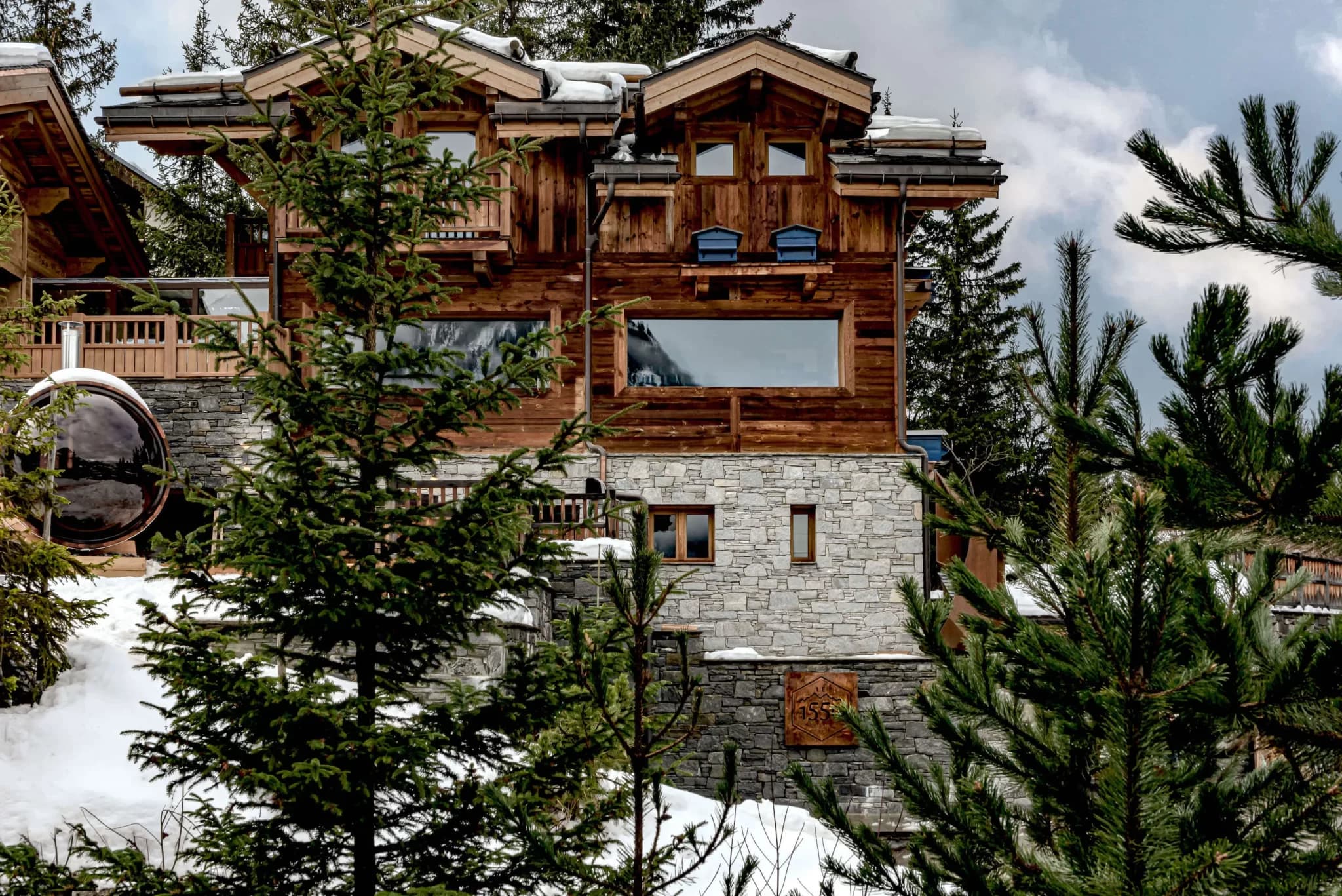Un luxueux chalet rustique avec un extérieur en bois et en pierre est niché dans un cadre de montagne enneigé et boisé en France. Des sapins entourent la maison, sur fond de premier plan enneigé et d'un ciel nuageux au-dessus, parfait pour une retraite de vacances inoubliable.