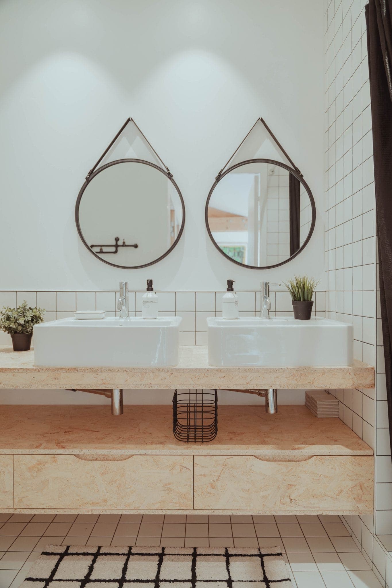 Modern bathroom with two white sinks on a wooden counter, round mirrors above each sink, small potted plants, soap dispensers, white tiled walls, and a black wire basket below the counter.