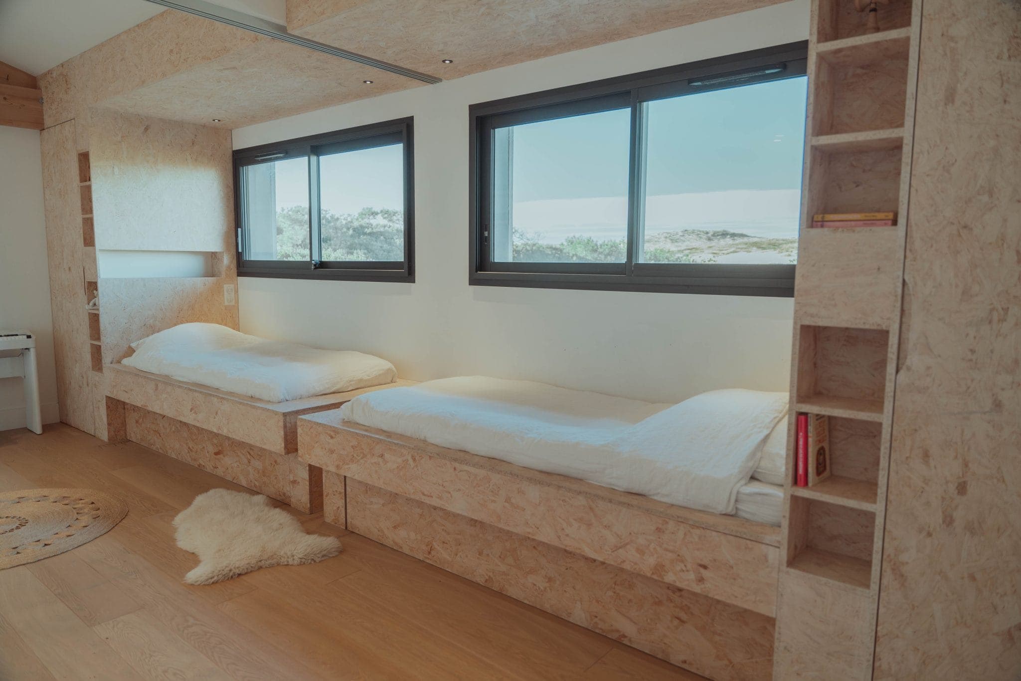 A minimalist bedroom with two single beds made of plywood, white bedding, large windows, built-in shelves, wooden floor, and a small white rug. Natural light fills the room.