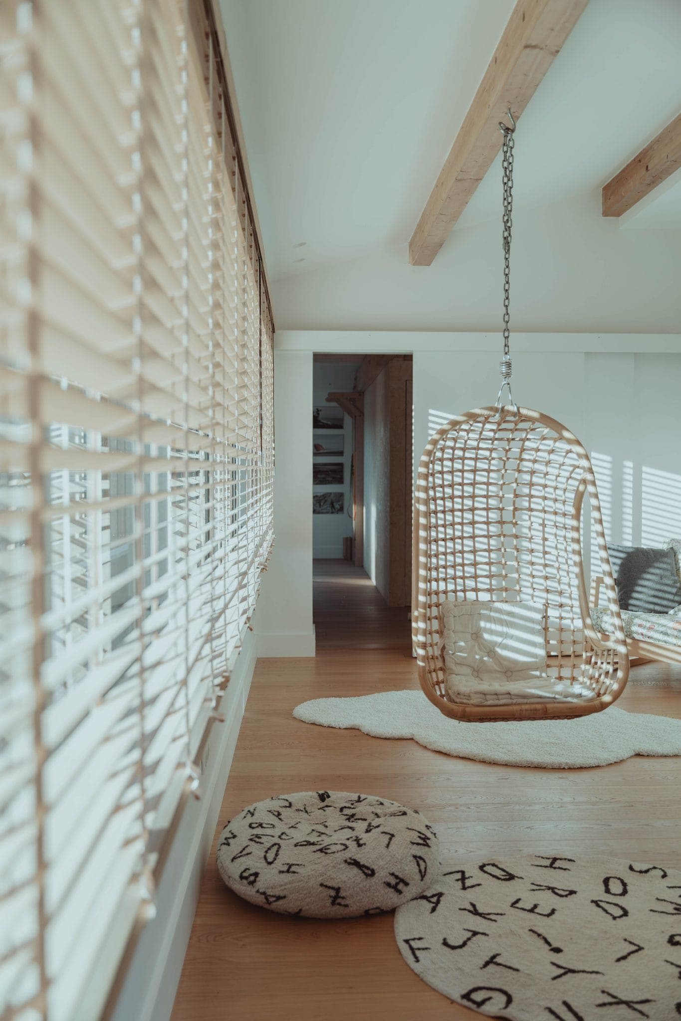 A bright room with wooden floors, large windows with blinds, a hanging rattan chair, white rugs, and round cushions with letters. Sunlight streams in, creating a cozy, airy atmosphere with natural decor.