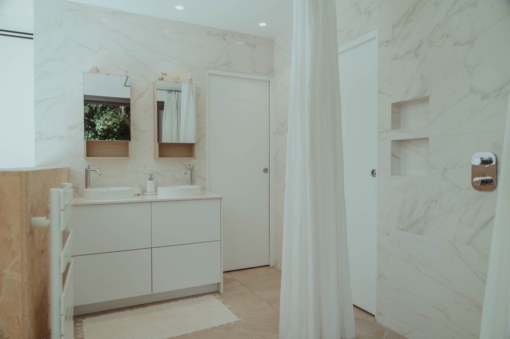 Modern bathroom with white marble walls, double sinks with mirrors, a white vanity, a rug, a shower area with a curtain, and built-in wall shelves. The space is bright and minimalistic.