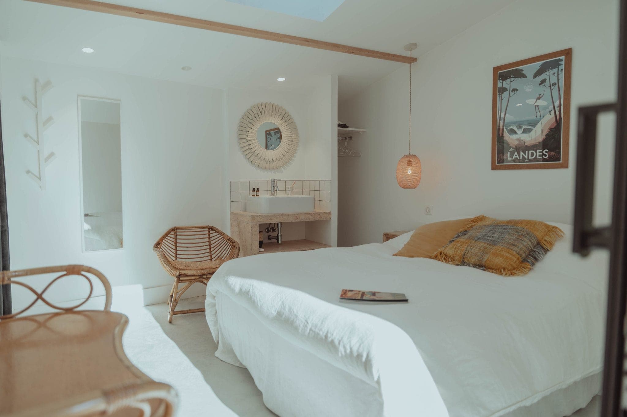 A cozy, sunlit bedroom with white walls and bedding, a wicker chair, a wooden desk, and a round mirror above a small sink. A pendant light and a framed "Landes" poster decorate the space. A magazine lies on the bed.