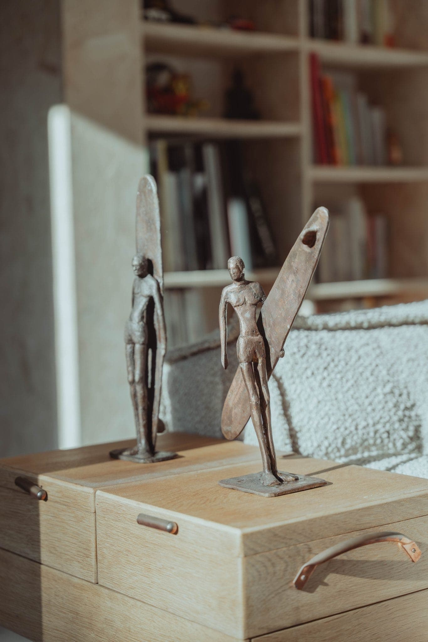 Two small metal sculptures of human figures holding surfboards stand on wooden drawers. In the background, there is a bookshelf with books and various items, softly lit by sunlight.