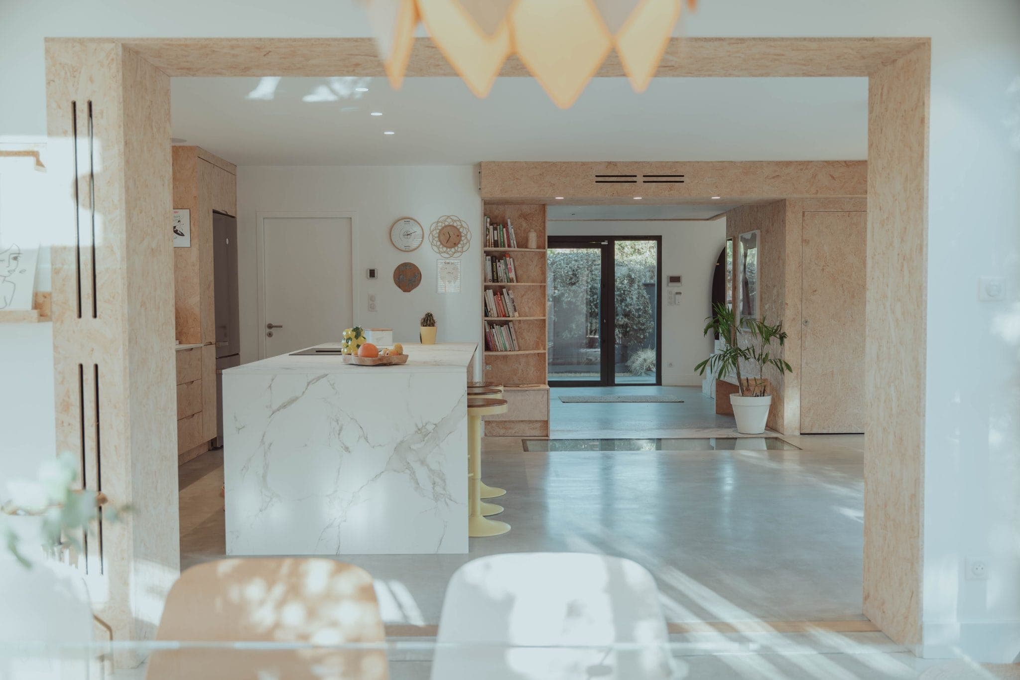 Modern, sunlit open-plan kitchen and living space with a marble island, wooden accents, houseplants, bookshelves, and large glass doors leading to a garden. Light and airy atmosphere with minimalist decor.