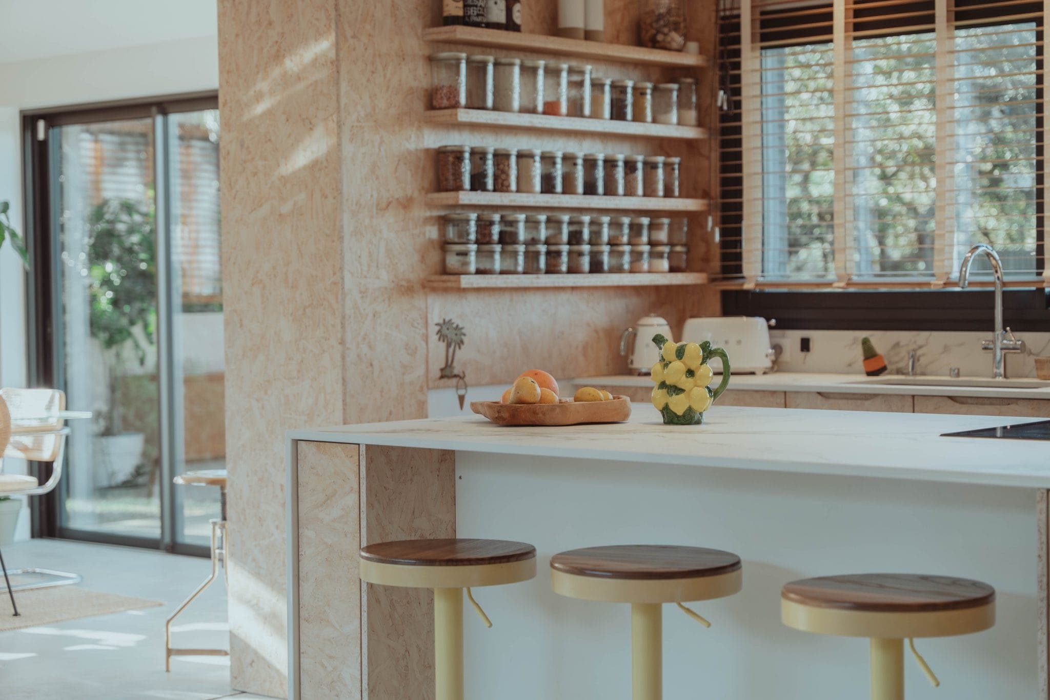 Modern kitchen with a white island, three wooden stools, a vase of yellow flowers, and a bowl of fruit. Shelves with spices, a window with blinds, and natural light fill the space.