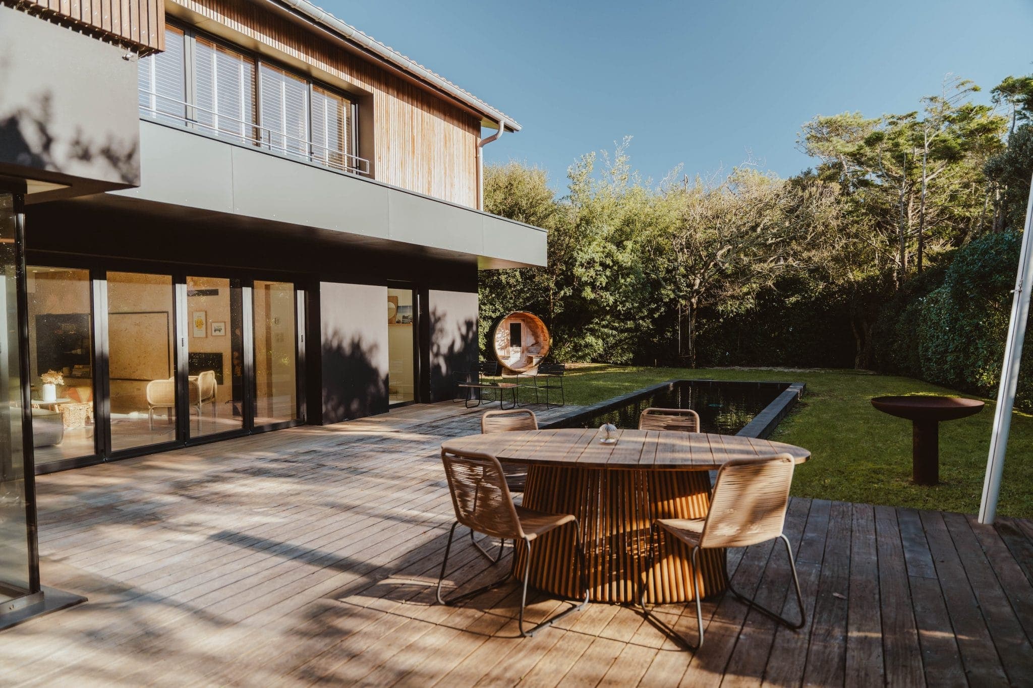 A modern house with large windows opens onto a wooden deck with a round table and chairs. The deck overlooks a narrow rectangular pool, surrounded by green lawn and trees under a clear blue sky.