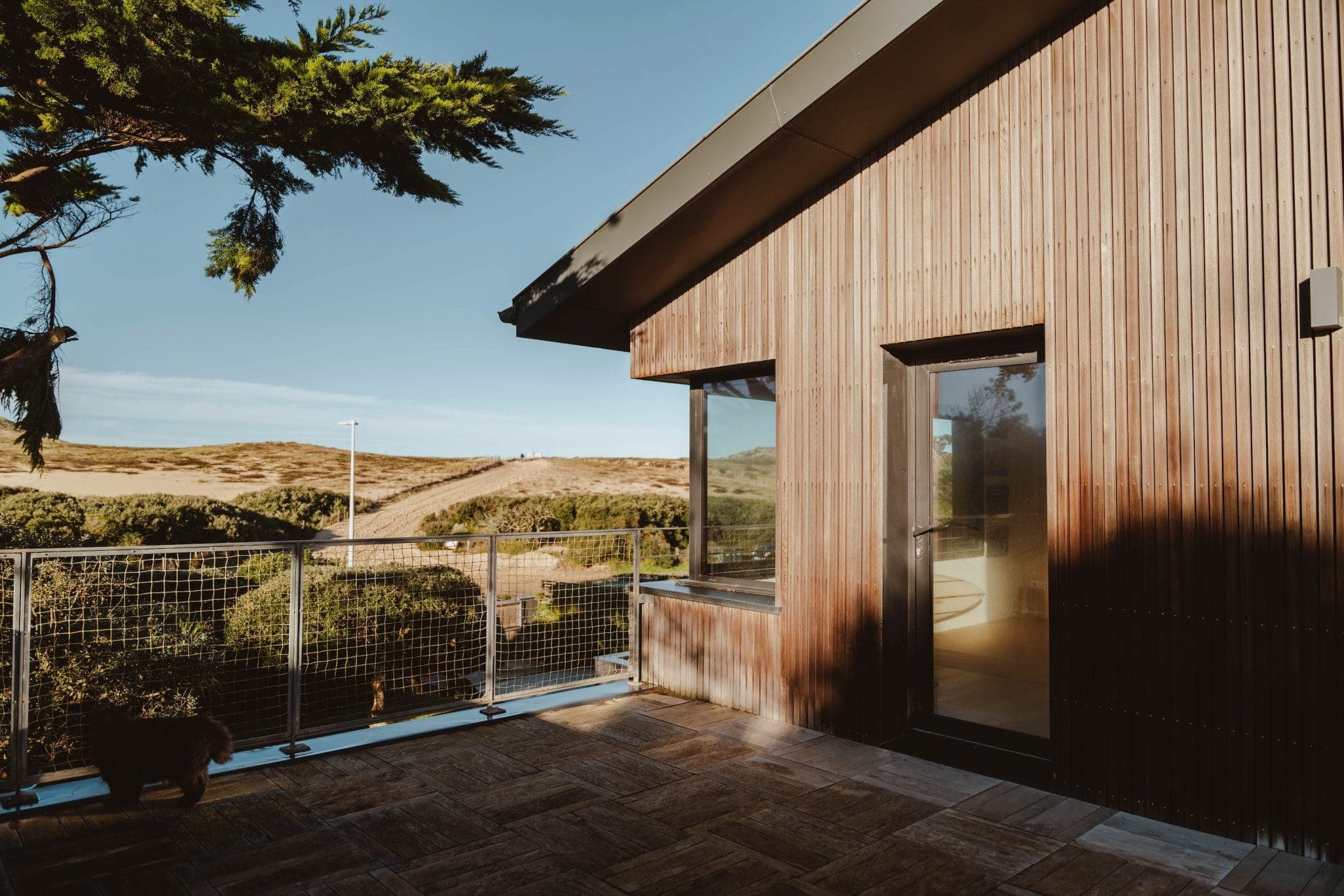 Une maison en bois moderne avec de grandes fenêtres et une porte vitrée s'ouvre sur un patio spacieux, donnant sur un paysage pittoresque de collines vallonnées et de verdure sous un ciel bleu clair.