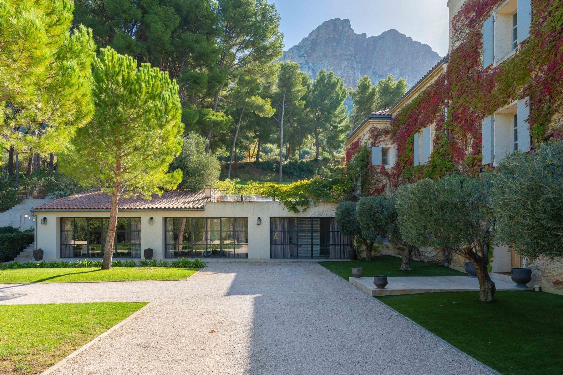 A picturesque villa surrounded by lush greenery. It features a traditional stone facade with vines, large windows, and a courtyard. Tall trees and a mountain peak are visible in the background under a clear blue sky.