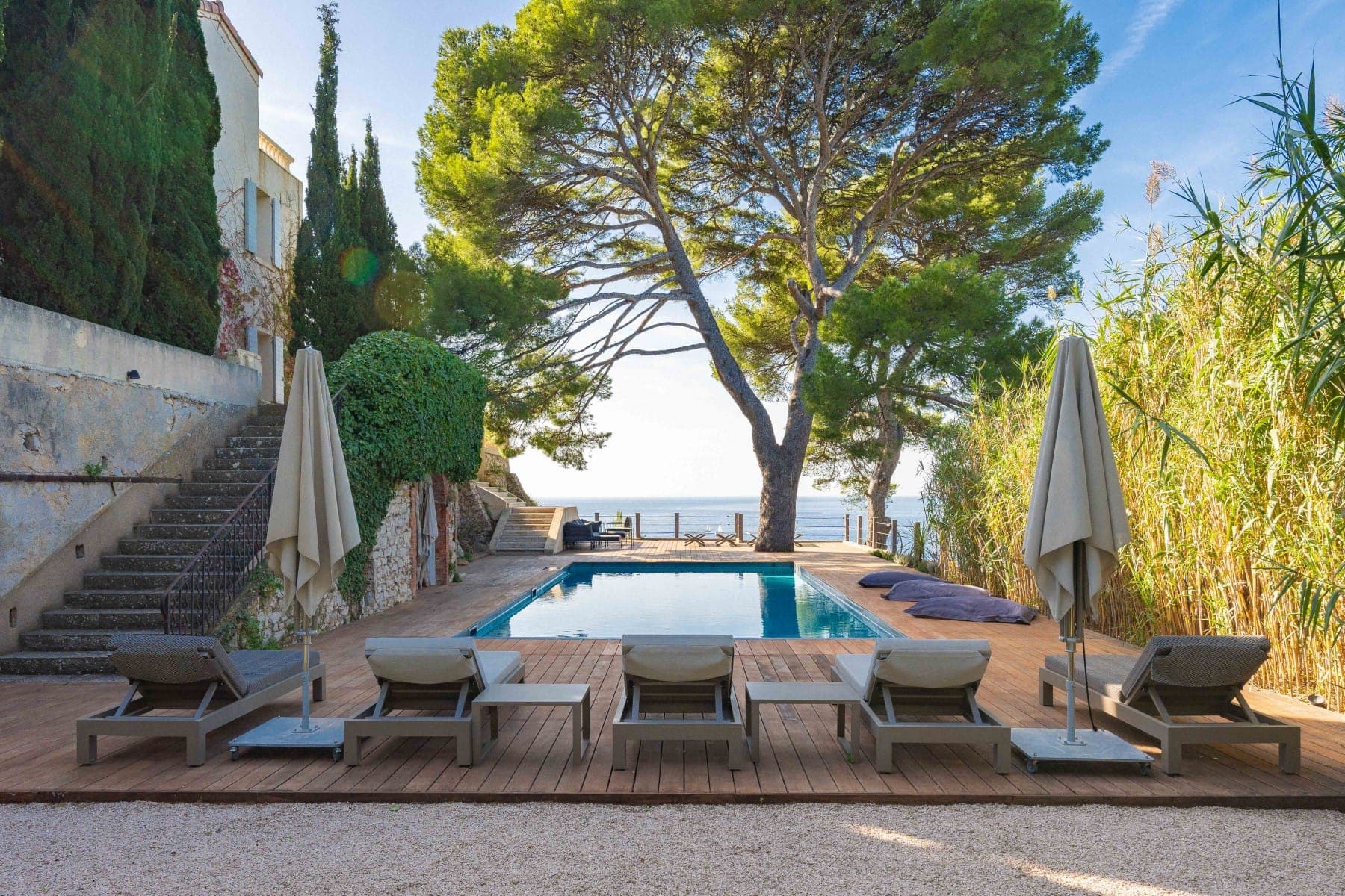 A serene poolside view featuring four lounge chairs and umbrellas on a wooden deck, overlooking a calm sea with a large tree in the background. Stairs and lush greenery surround the area, creating a tranquil, inviting atmosphere.