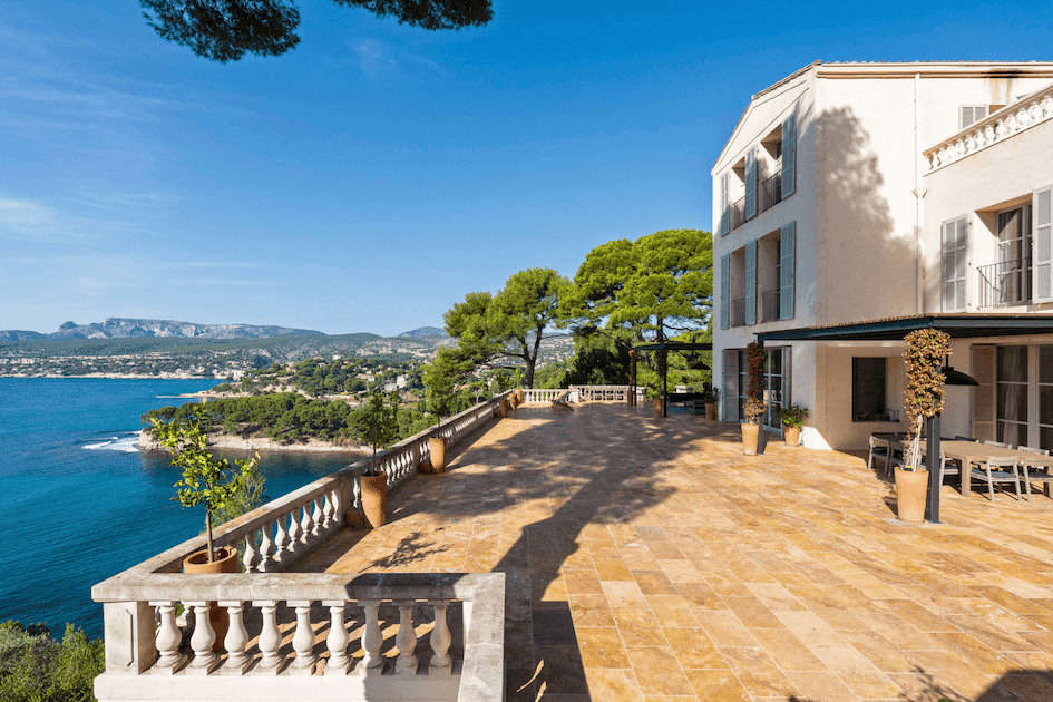 A spacious terrace with tan stone tiles overlooks a coastal landscape. The view includes lush greenery, a calm blue sea, and distant mountains. A white building with tall windows and potted plants lines the right side of the terrace.