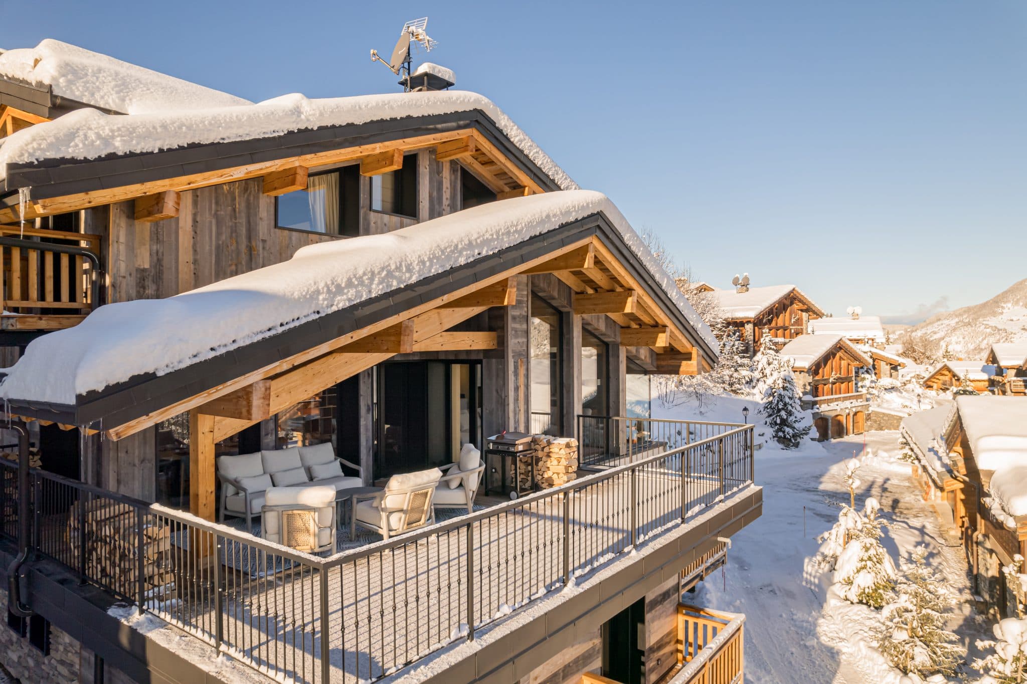 Un chalet de montagne confortable à Courchevel avec des toits enneigés et un balcon ensoleillé avec des sièges extérieurs. Les environs sont recouverts de neige et d'autres chalets peuvent être vus en arrière-plan sous un ciel bleu clair.