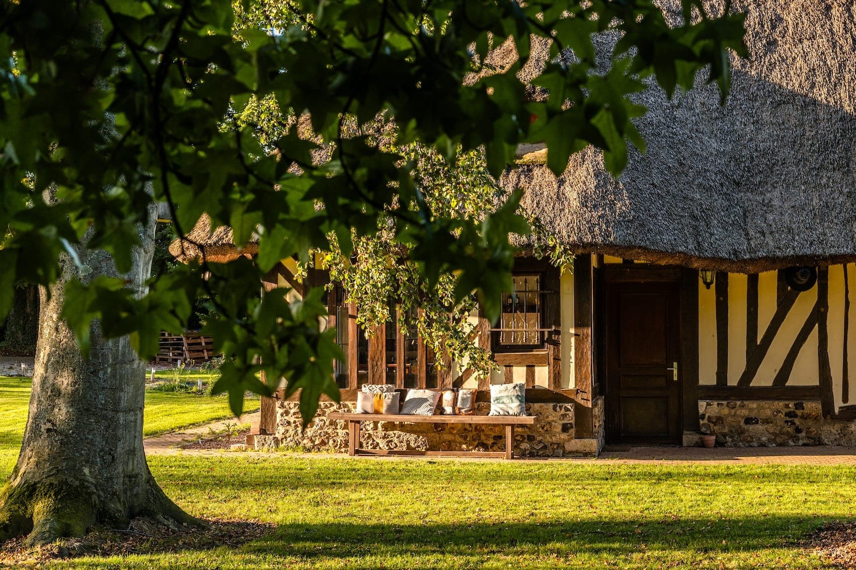 A cozy cottage with a thatched roof and wooden beams, reminiscent of Normandie charm, is partially obscured by tree leaves in the foreground. A wooden bench with cushions sits under a window on a sunny lawn, creating a peaceful and inviting atmosphere.
