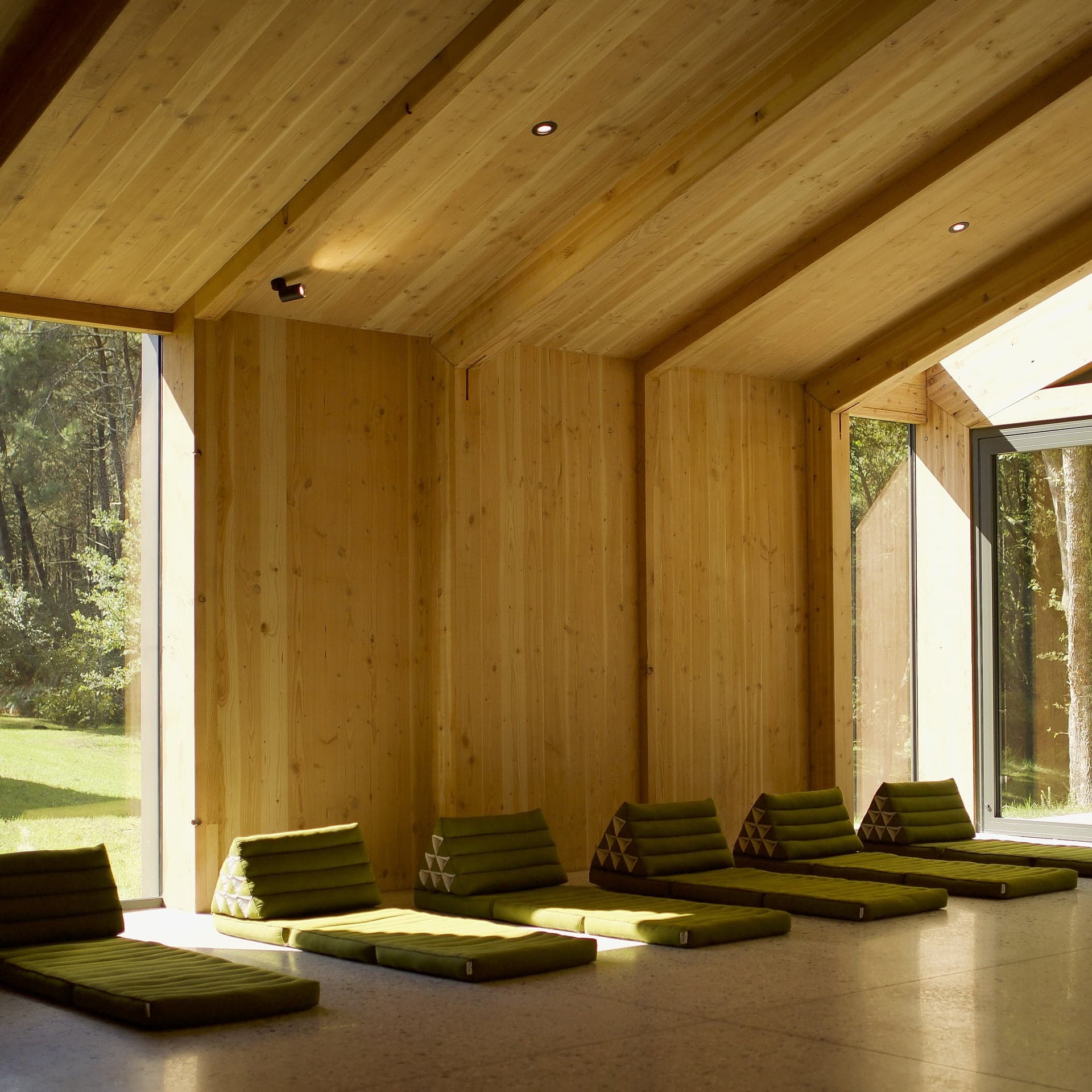Un lieu de retraite serein avec des murs et des plafonds en bois inclinés, avec une rangée de chaises longues vertes rembourrées. De grandes fenêtres encadrent la forêt ensoleillée à l'extérieur, parfaite pour le yoga au milieu de l'étreinte tranquille de la nature dans cet espace paisible et minimaliste.