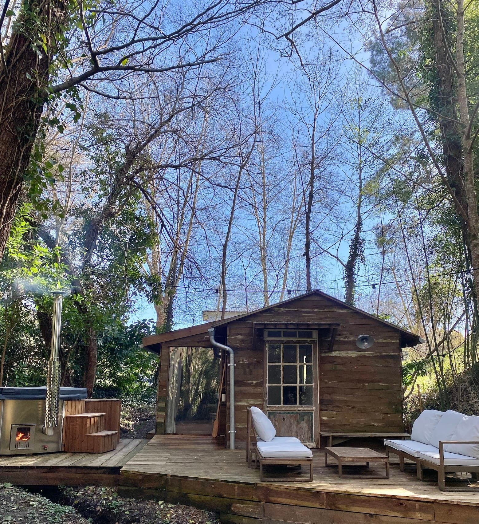 Une cabane rustique en bois avec une terrasse, des sièges extérieurs et un jacuzzi est entourée de grands arbres sans feuilles sous un ciel bleu clair. De la vapeur s'échappe du jacuzzi, créant une atmosphère chaleureuse.