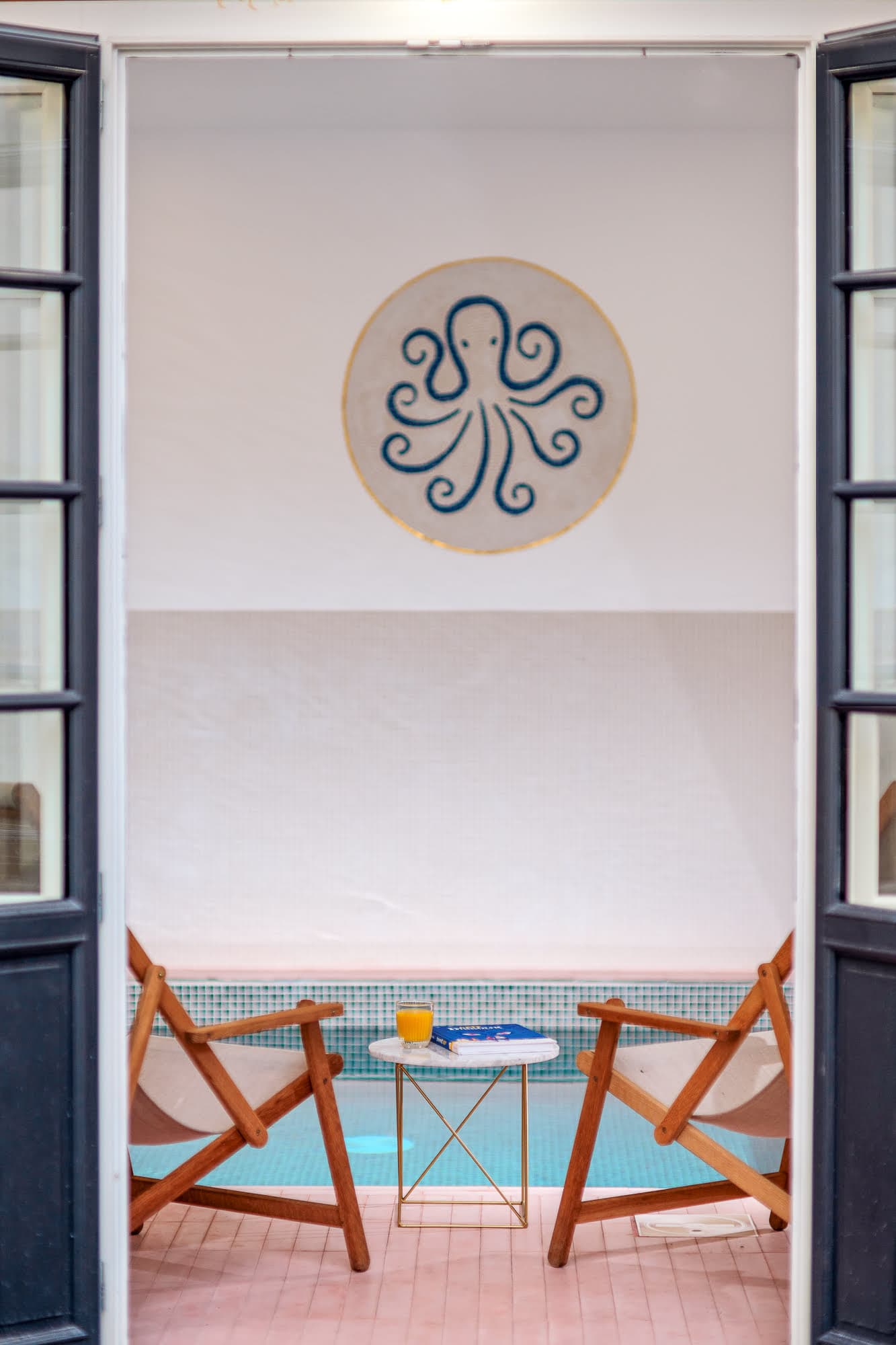 Vue d'un espace piscine intérieur cosy dans un endroit bord de mer en Normandie, avec des portes ouvertes révélant deux chaises en bois faisant face à une petite table ronde avec un verre de jus d'orange. Le mur arbore un impressionnant motif de pieuvre, reflétant parfaitement l'ambiance maison vacances.