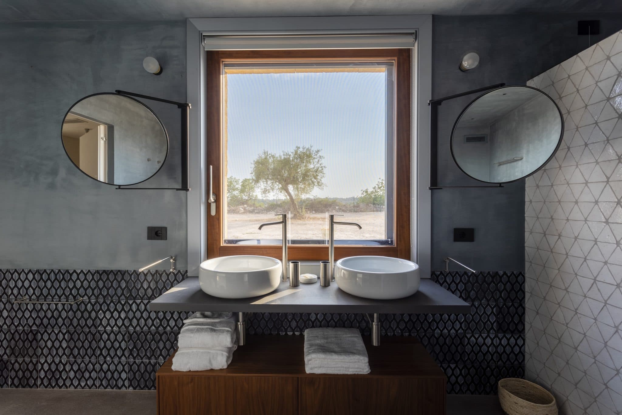 A modern bathroom with a large window featuring an outdoor view of a tree and a clear sky. Two round mirrors are mounted on either side of the window above two white vessel sinks, with a wooden cabinet below holding towels. The wall features a geometric tile design.