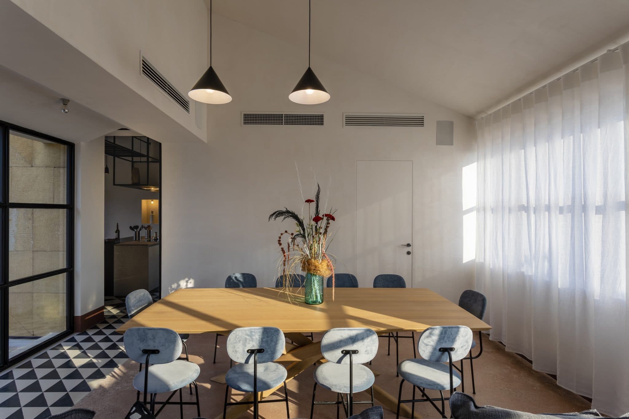A modern dining room with a large wooden table surrounded by eight cushioned chairs. Two pendant lights hang above the table. Natural light shines through sheer white curtains, illuminating the space. A vase with dried plants and flowers sits in the center of the table.