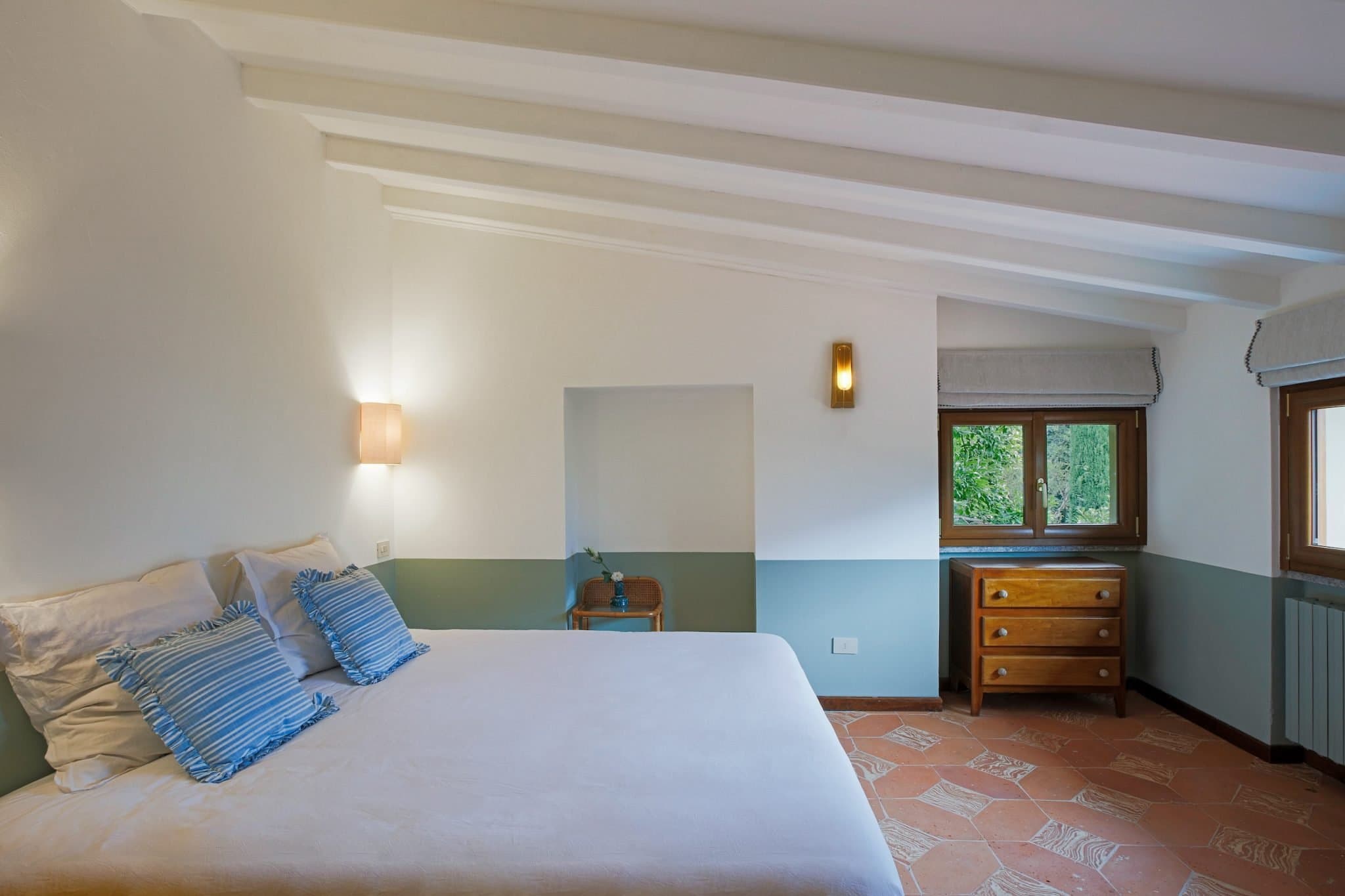 A cozy bedroom with a sloped ceiling, featuring a neatly made bed with white sheets and blue accent pillows, a small wooden desk with a chair, a dresser with a window above it, and terracotta tile flooring. The room has a tranquil and simple decor.