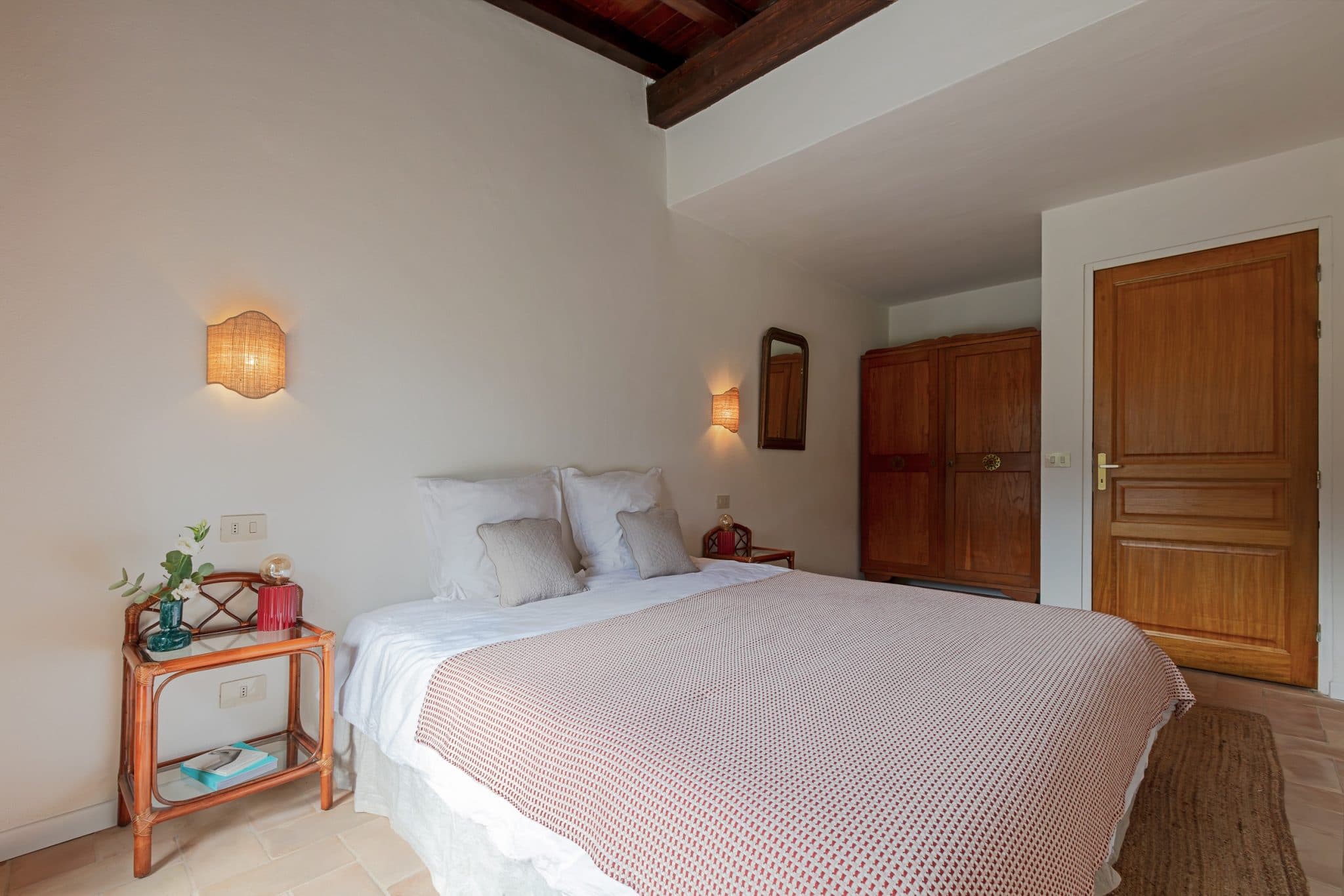 Image of a cozy bedroom with a large wooden bed covered with white and pink bedding. There are light fixtures and small bedside tables with decor on either side. A wooden wardrobe and a door are visible in the background. The room has white walls and a wooden ceiling.