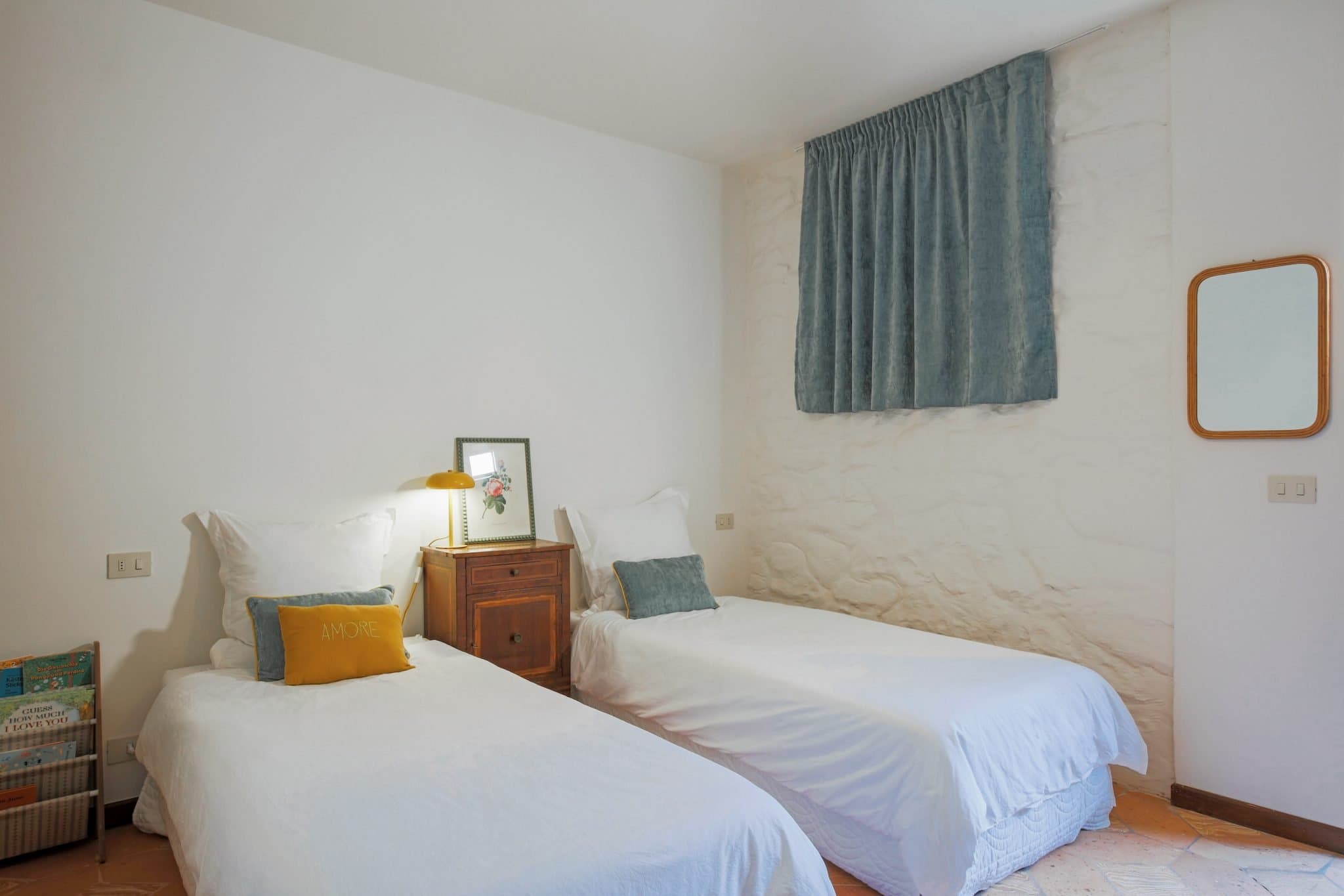 A simple, well-lit bedroom with two single beds covered in white bedspreads and accented with decorative pillows. A small wooden nightstand with a lamp, framed picture, and books stands between the beds. A mirror hangs on a wall, and a window is covered with a blue curtain.