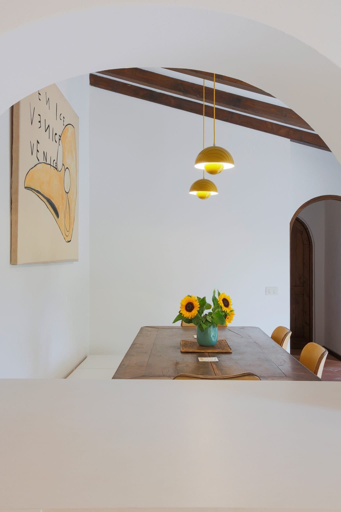 A cozy dining area with a wooden table set for four, featuring a vase of sunflowers at its center. Three modern, gold pendant lights hang above the table. An archway frames the view, and a piece of abstract art decorates the white wall behind the table.