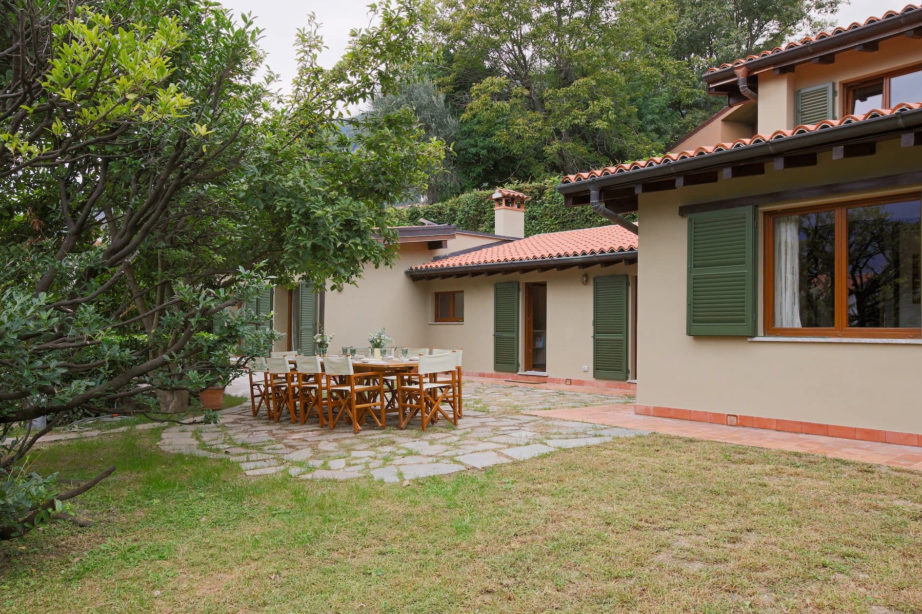 A charming house with light-colored walls and green shutters features a spacious patio with a large outdoor dining table and chairs. The garden is lush with green grass, trees, and bushes, creating a serene and inviting atmosphere.