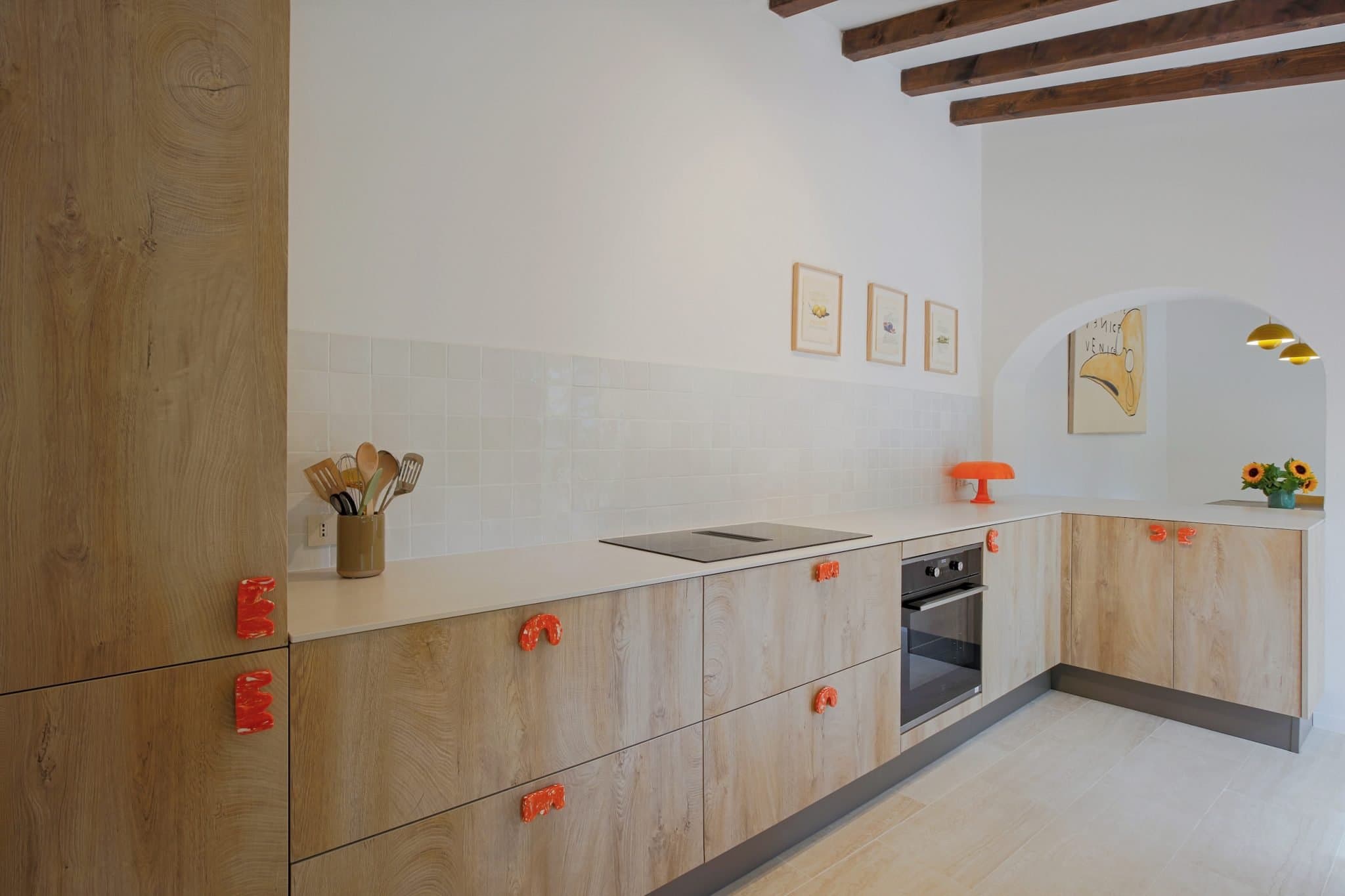 A modern kitchen with wooden cabinets and a white tiled backsplash. The oven and stovetop are built into the counter. Bright orange handles adorn the cabinets, and on the countertop are a utensil holder and an orange lamp. Sunflowers in a vase are on the right.