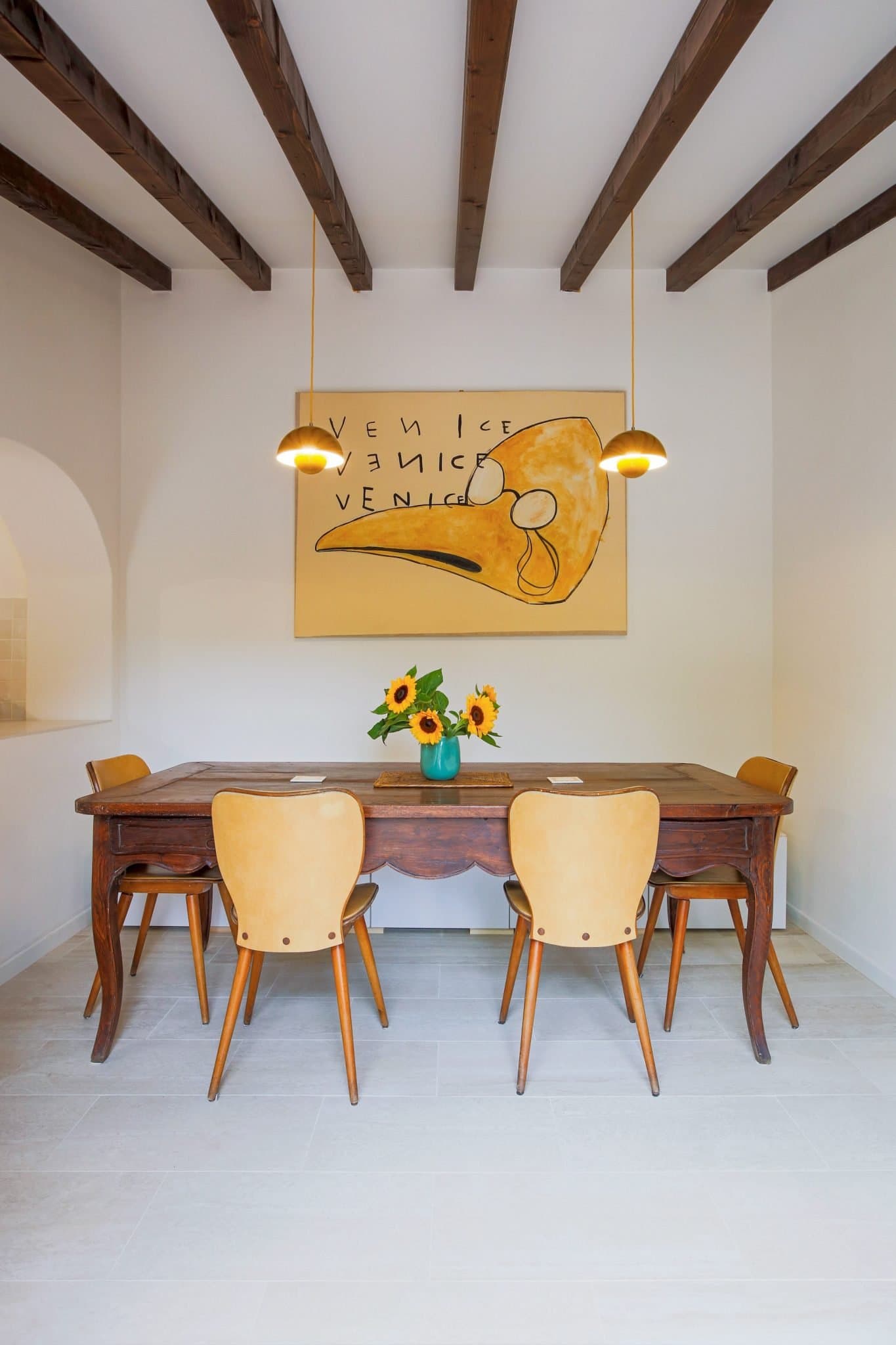 A cozy dining room with a wooden table and six matching chairs. Above the table hang two pendant lights, and a vase with sunflowers sits at the center. The room features exposed wooden ceiling beams and a large wall art piece with the word "Venice.