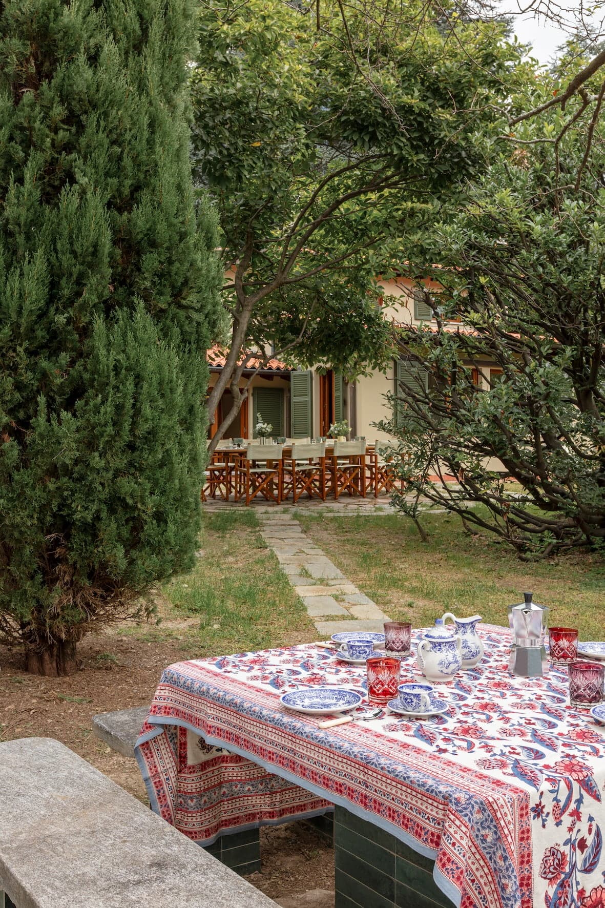 A picnic setup with a colorful tablecloth, tea set, and cups on a stone table is in the foreground, beneath a tree. In the background, there's a pathway leading to a house with a patio that has wooden chairs and tables in a garden setting.