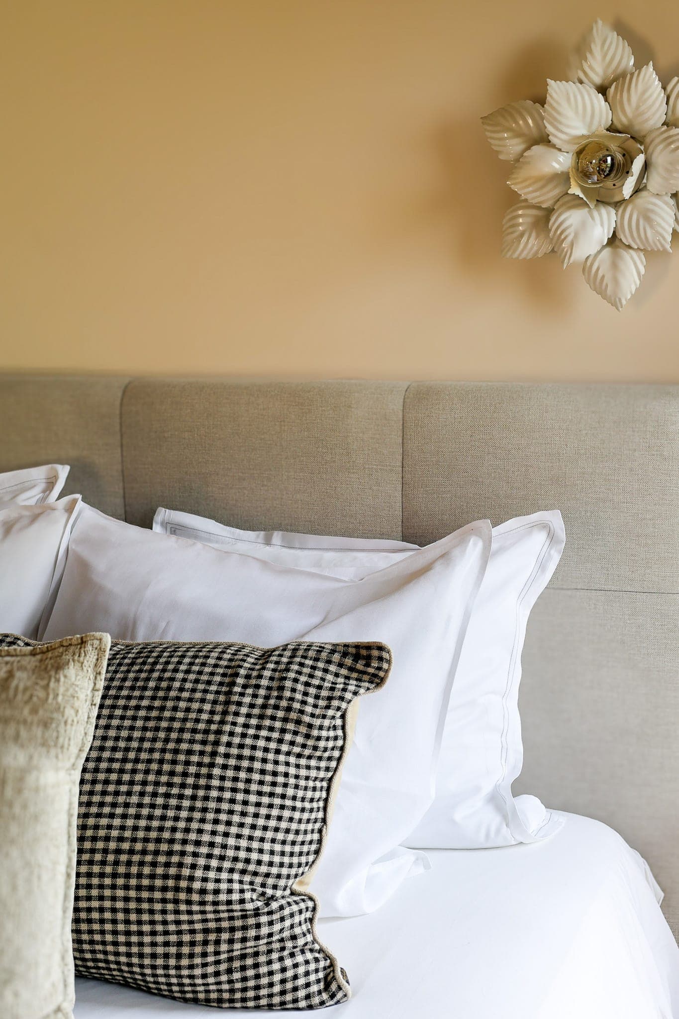 Un lit impeccablement fait avec des oreillers blancs et un coussin à carreaux noirs et blancs, posé sur une tête de lit beige clair. Une décoration murale florale blanche est visible à droite de l'image.