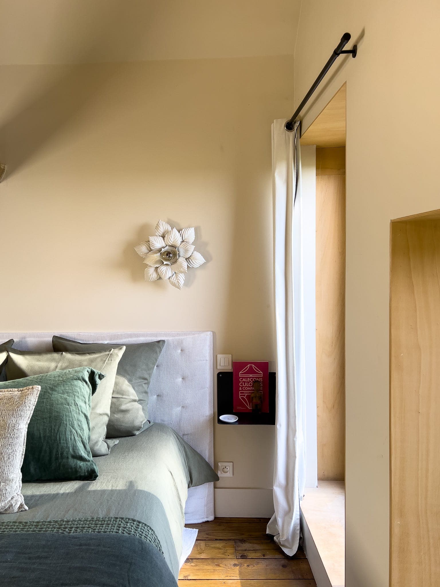 Un coin chambre cosy avec un lit vert, une tête de lit blanche, des coussins décoratifs et une décoration murale à fleurs blanches. La lumière pénètre par une fenêtre aux rideaux blancs, et un livre est posé sur une petite étagère murale à proximité.
