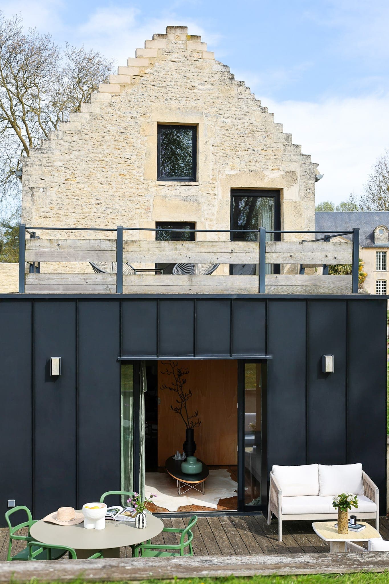 Une maison moderne avec un étage supérieur en pierre et un étage inférieur en métal noir, dotée de grandes fenêtres, d'une terrasse sur le toit avec des sièges et d'un patio avec table, chaises et canapé d'extérieur. Des arbres sont visibles en arrière-plan.