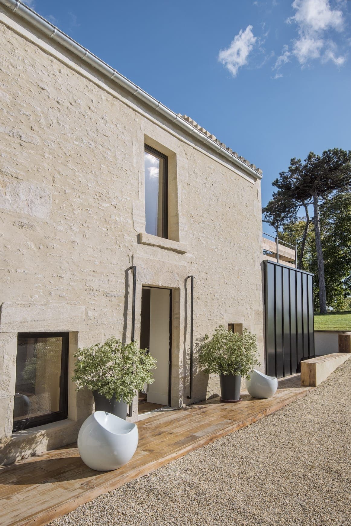 Une maison en pierre moderne avec de hautes fenêtres étroites, une terrasse en bois, de grandes plantes en pot et une extension en métal noir, sur fond d'arbres et d'un ciel bleu avec quelques nuages.