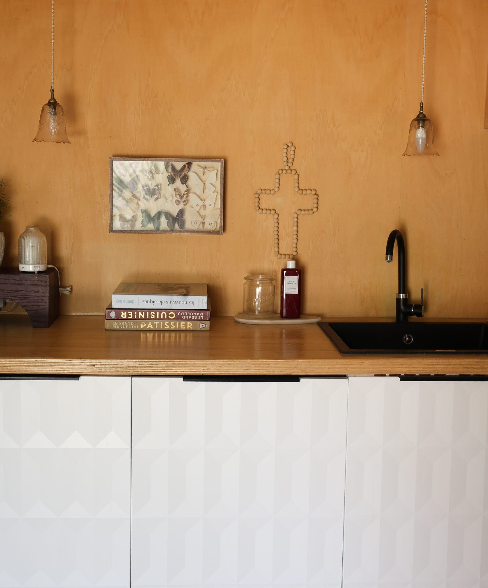 Minimalist kitchen with wooden wall, black sink, books, and a glass jar on the counter. Two pendant lights hang above, with a framed picture and decorative cross on the wall. White geometric cabinets below.