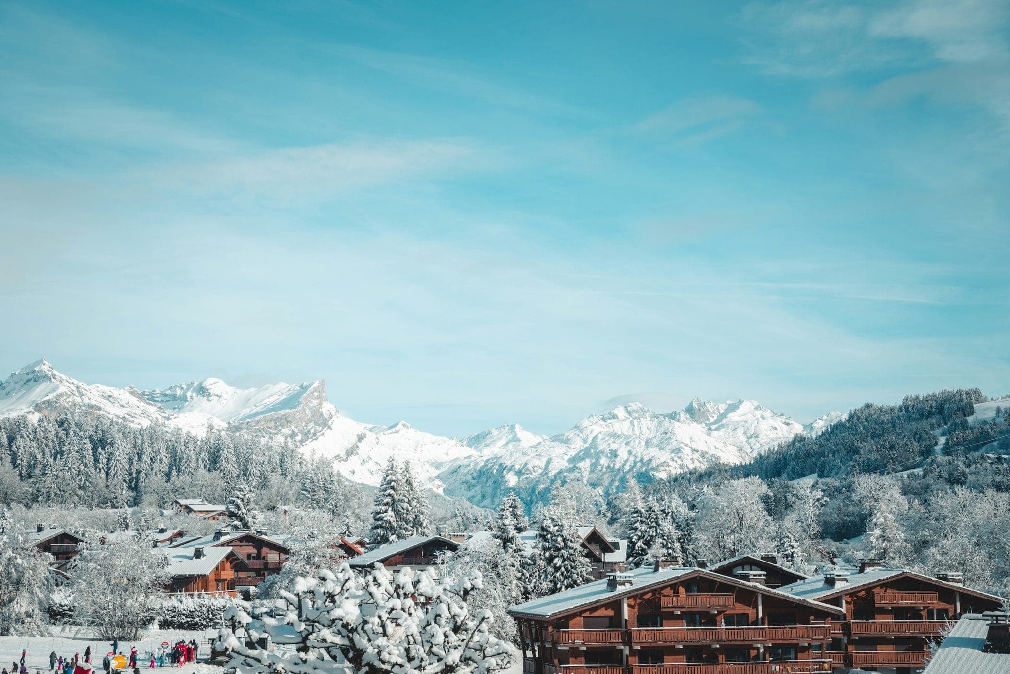 Village de montagne enneigé avec ses chalets en bois nichés au milieu des arbres enneigés. De majestueuses montagnes s'élèvent en arrière-plan sous un ciel bleu azur. Au loin, on aperçoit des skieurs et des touristes vêtus de tenues colorées.