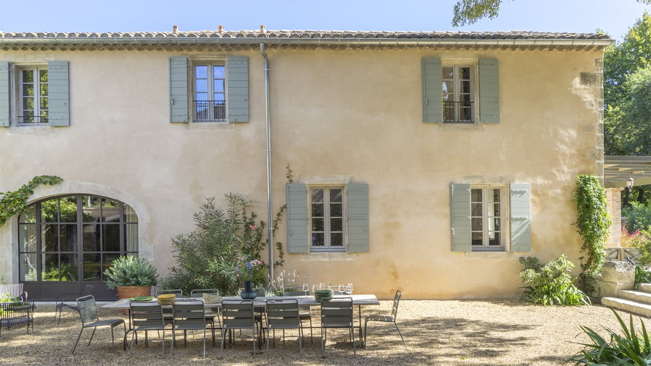 Une maison rustique à deux étages avec des murs en stuc beige et des volets vert sauge. Une longue table à manger extérieure et des chaises sont installées sur un patio en gravier au premier plan. La scène est entourée de verdure et baignée de soleil, suggérant une journée paisible et chaude.