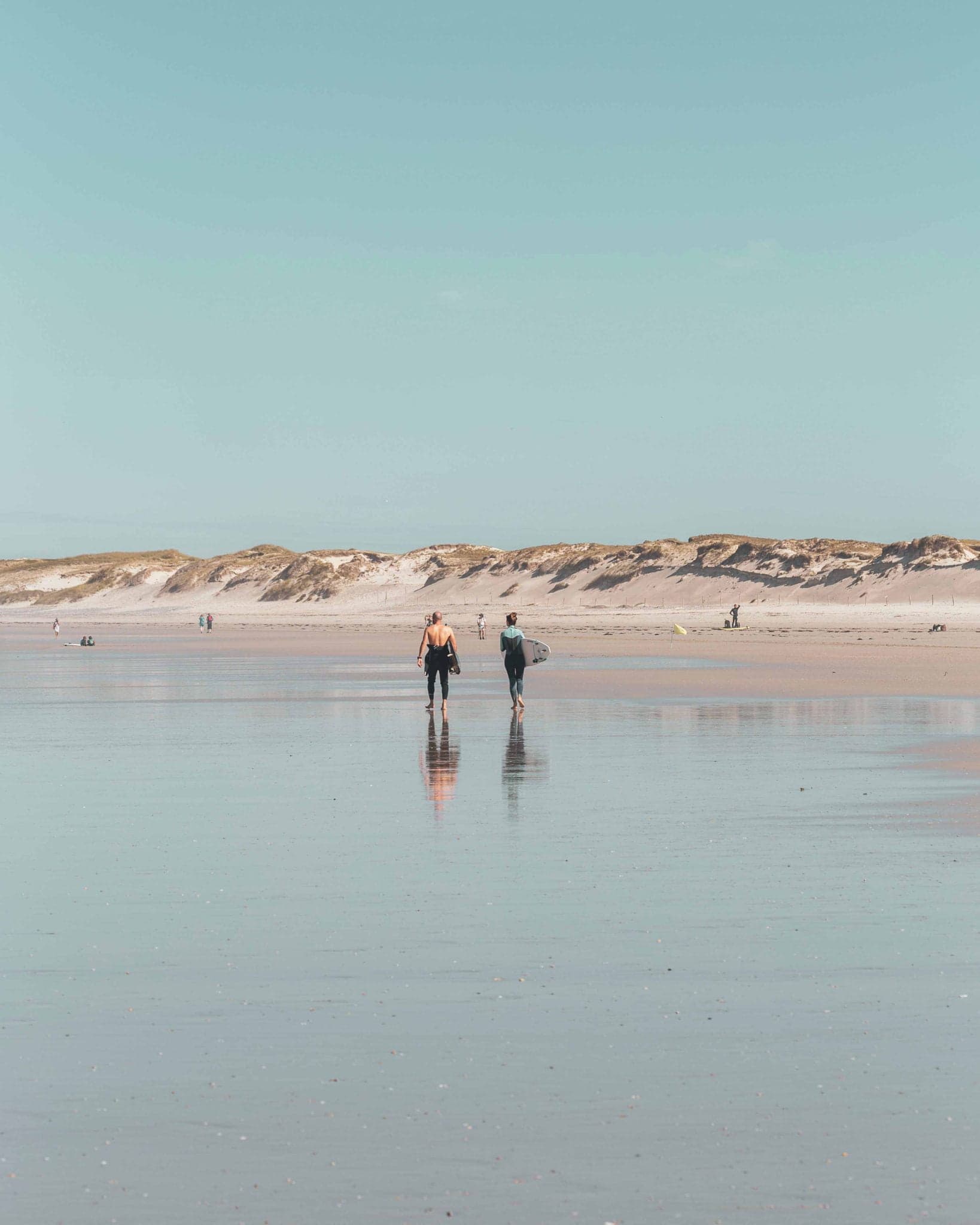 Deux surfeurs marchant sur la plage, dunes et ciel bleu en arrière plan