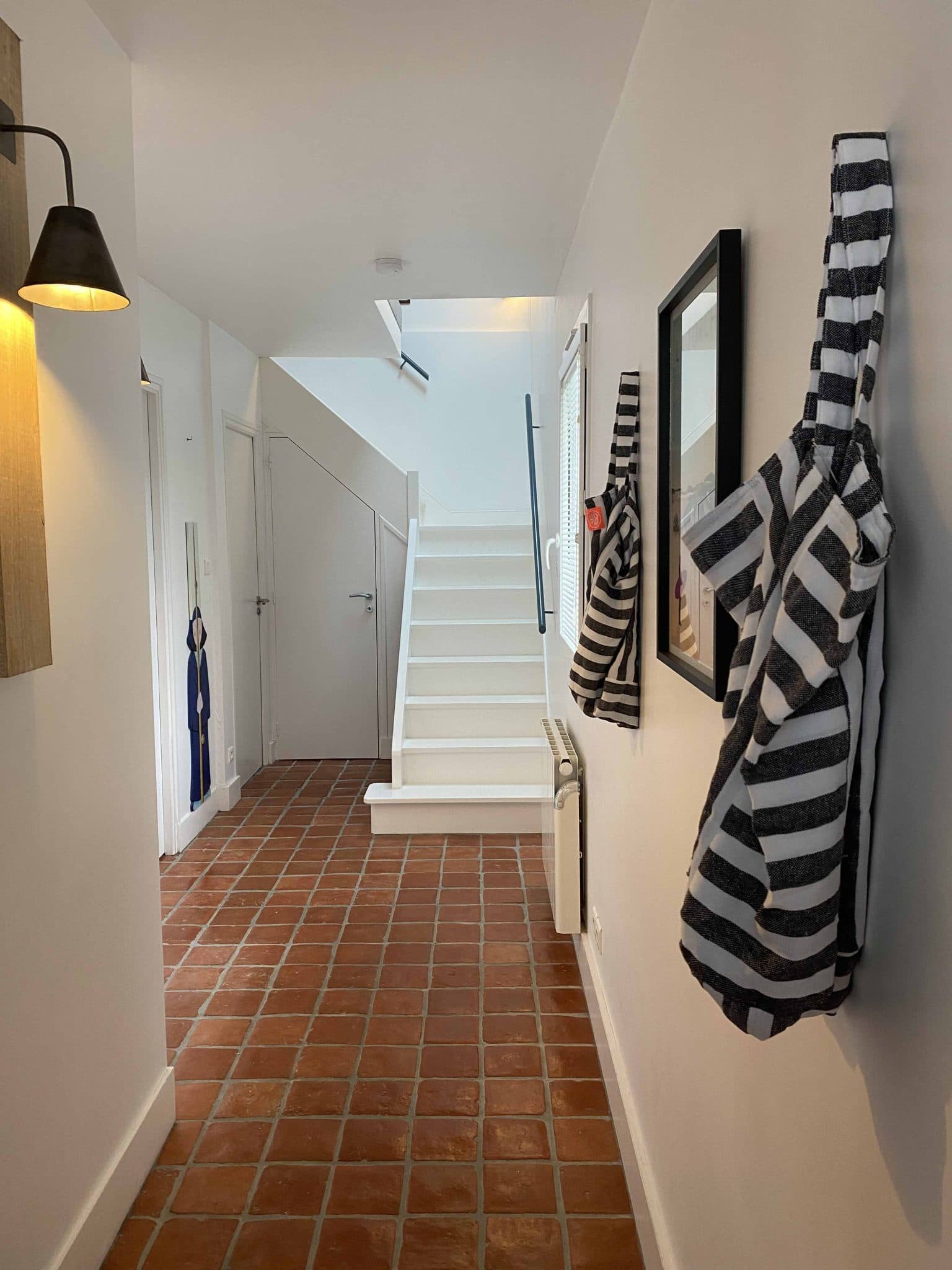 Couloir avec carrelage au sol, escalier blanc et cirés accrochés au porte-manteau