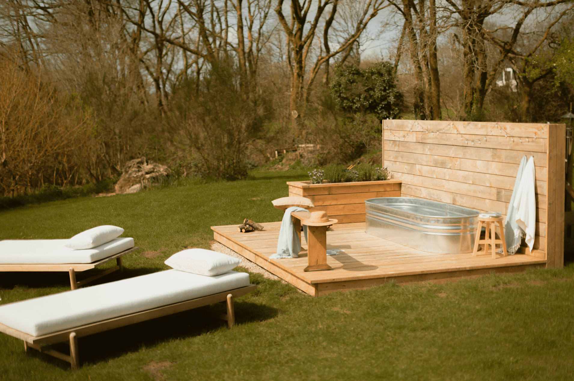Une terrasse extérieure en bois avec une baignoire en métal, une petite table et deux chaises longues à coussins blancs sur l'herbe, entourée d'arbres et de verdure par une journée ensoleillée.