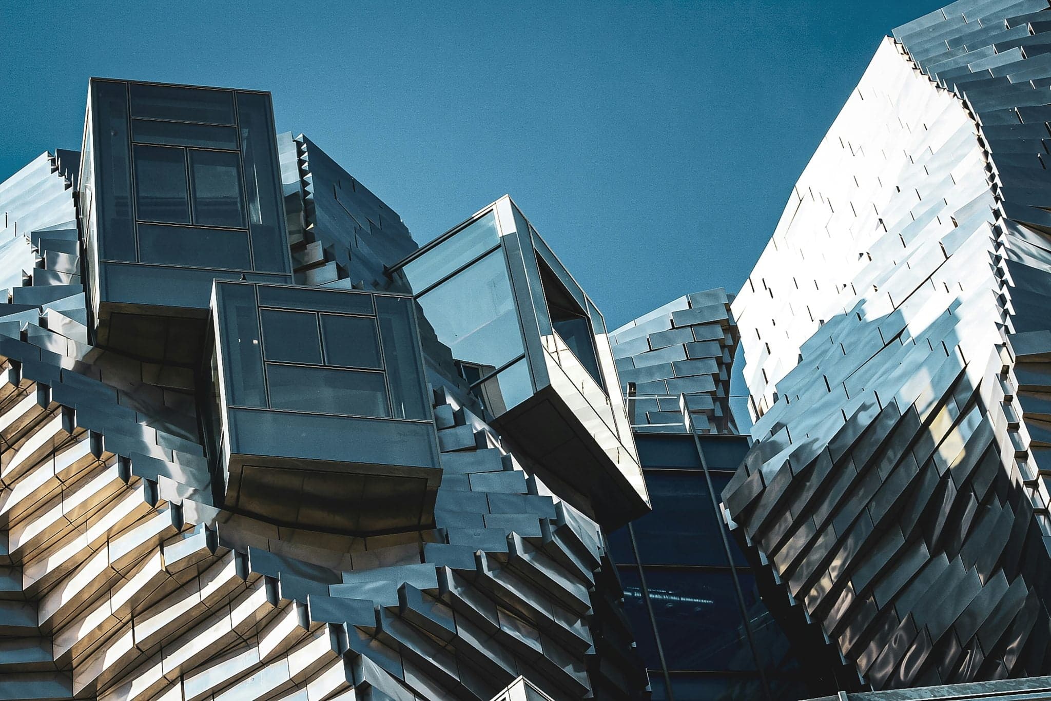 Ce bâtiment futuriste d'Arles présente un design angulaire saisissant avec des surfaces métalliques réfléchissantes et des fenêtres rectangulaires en saillie sur fond de ciel bleu clair.