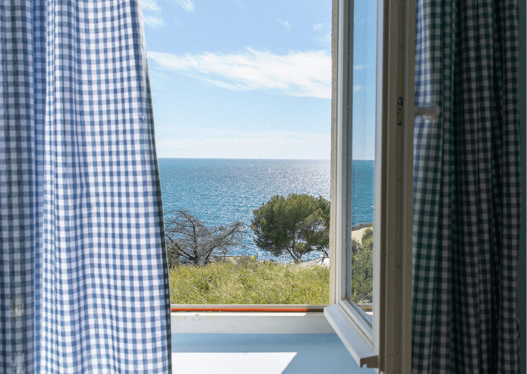 Admirez la vue sereine depuis une villa de Marseille à travers une fenêtre ouverte ornée de rideaux à carreaux bleus et blancs. Admirez le paysage océanique pittoresque, encadré par des arbres et des champs herbeux sous un ciel bleu clair.
