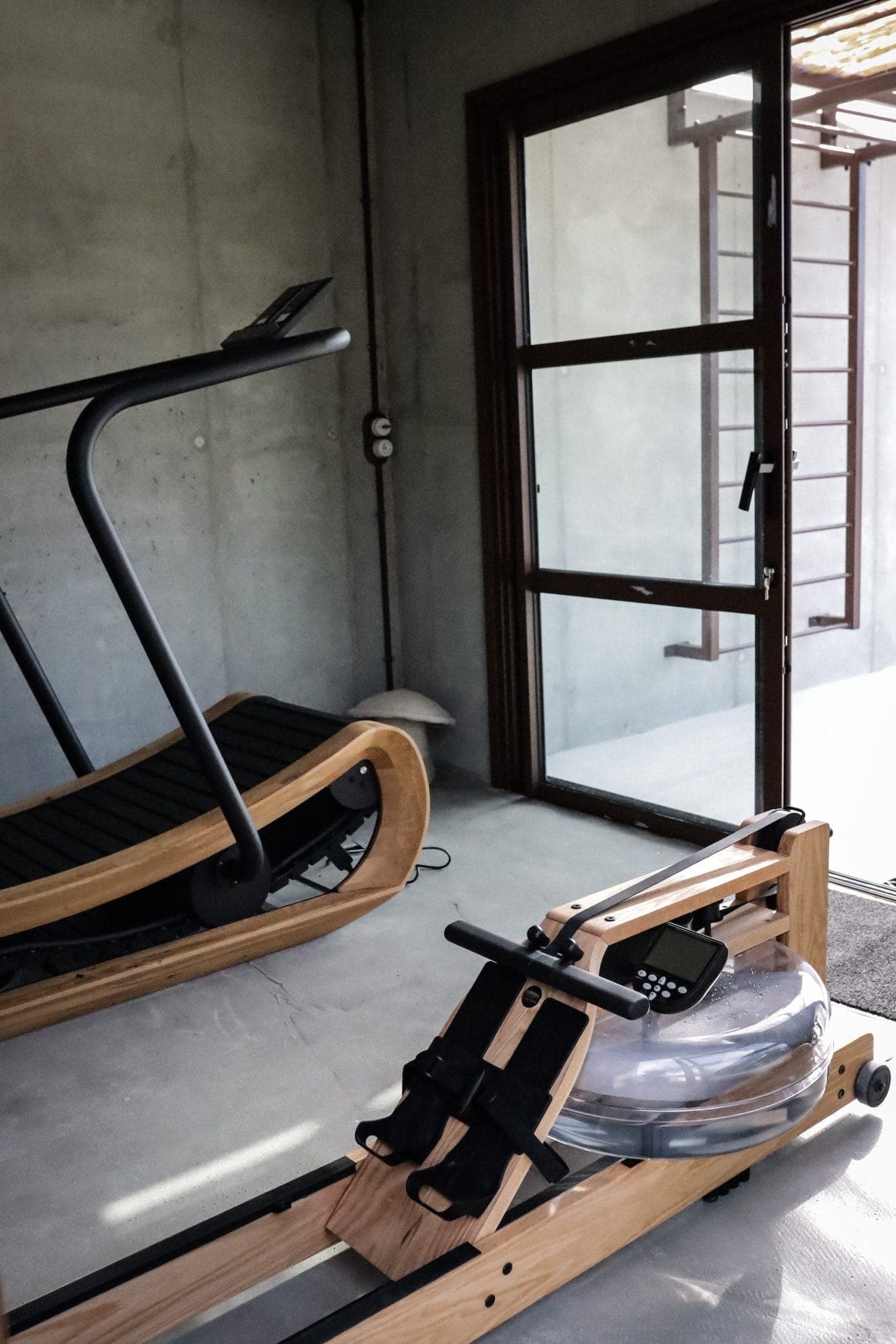 A minimalist gym with a wooden treadmill and rowing machine on a concrete floor. Large glass doors let in natural light and an outside ladder is visible through the doors. The interior has an elegant, modern design.