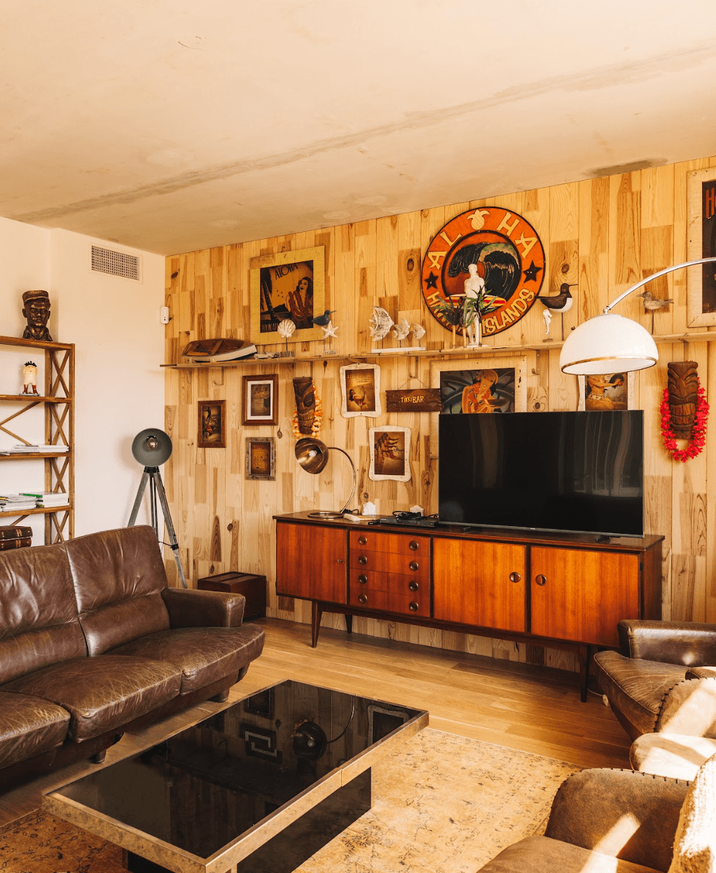 Un salon confortable avec des murs en bois ornés de diverses décorations vintage, telles que des tableaux encadrés, des masques et une pancarte hawaïenne. La chambre comprend un grand canapé en cuir marron, une table basse au dessus noir, une télévision à écran plat sur une console en bois et un lampadaire.