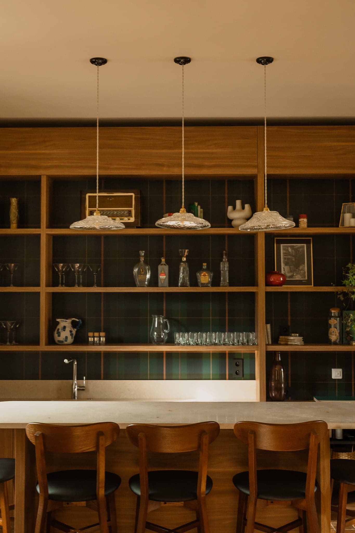Une cuisine confortable avec des armoires en bois, trois suspensions, un comptoir blanc et quatre chaises en bois. Les étagères en arrière-plan présentent divers objets, notamment des bouteilles, des verres, des objets de décoration et une radio vintage.