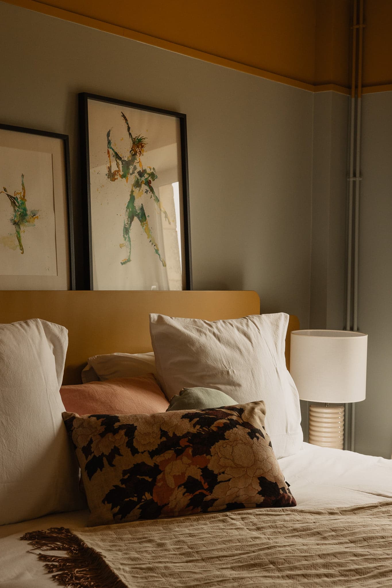 Une chambre confortable comprenant un lit avec plusieurs oreillers de différentes couleurs, dont du blanc, du pêche et un à motifs floraux. Il y a une couverture blanc cassé drapée sur le lit. Derrière le lit se trouvent deux œuvres d'art abstraites encadrées sur le mur. Une lampe est posée sur une table de chevet.