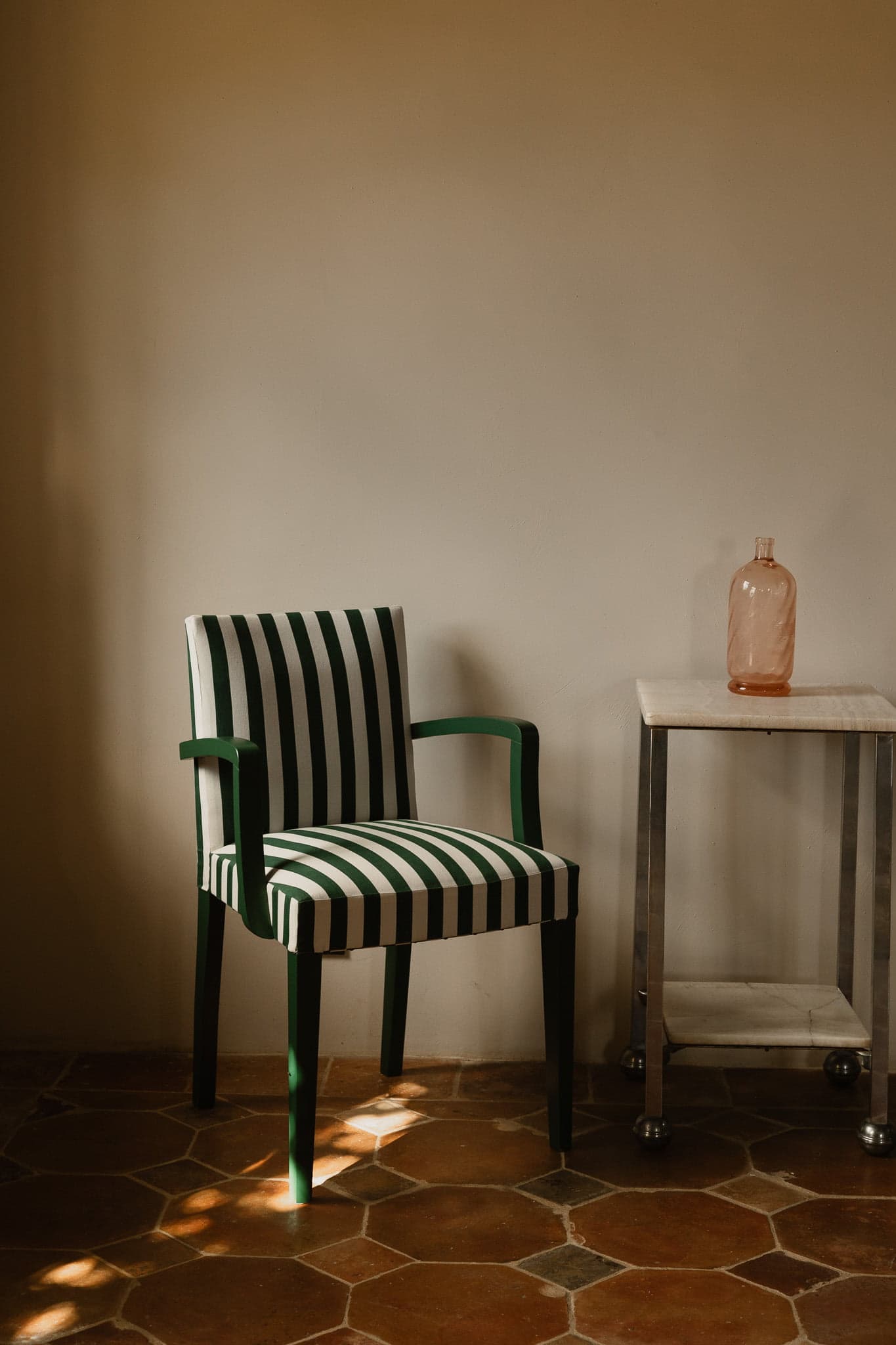 Un élégant fauteuil à rayures vertes et blanches trône à côté d'une petite table en métal et en marbre. La table contient une seule bouteille en verre rose translucide. Les deux objets sont posés sur un sol carrelé avec une lumière douce et naturelle projetant des ombres.