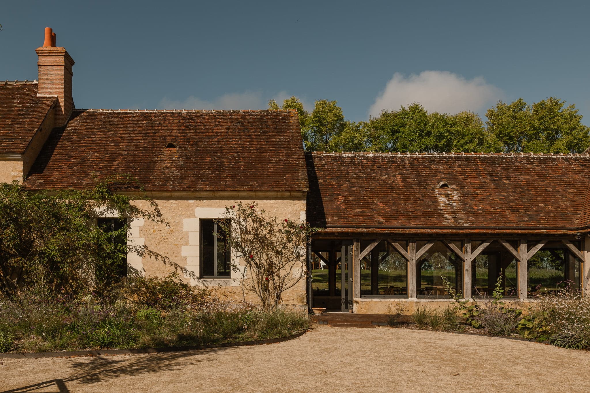 Un bâtiment rustique en pierre avec un toit de tuiles rouges et des poutres en bois, bordé par une allée sablonneuse et un espace vert paysager. De grandes fenêtres révèlent l'intérieur, entouré de plantes à fleurs. En arrière-plan, de grands arbres se dressent sur un ciel bleu clair.