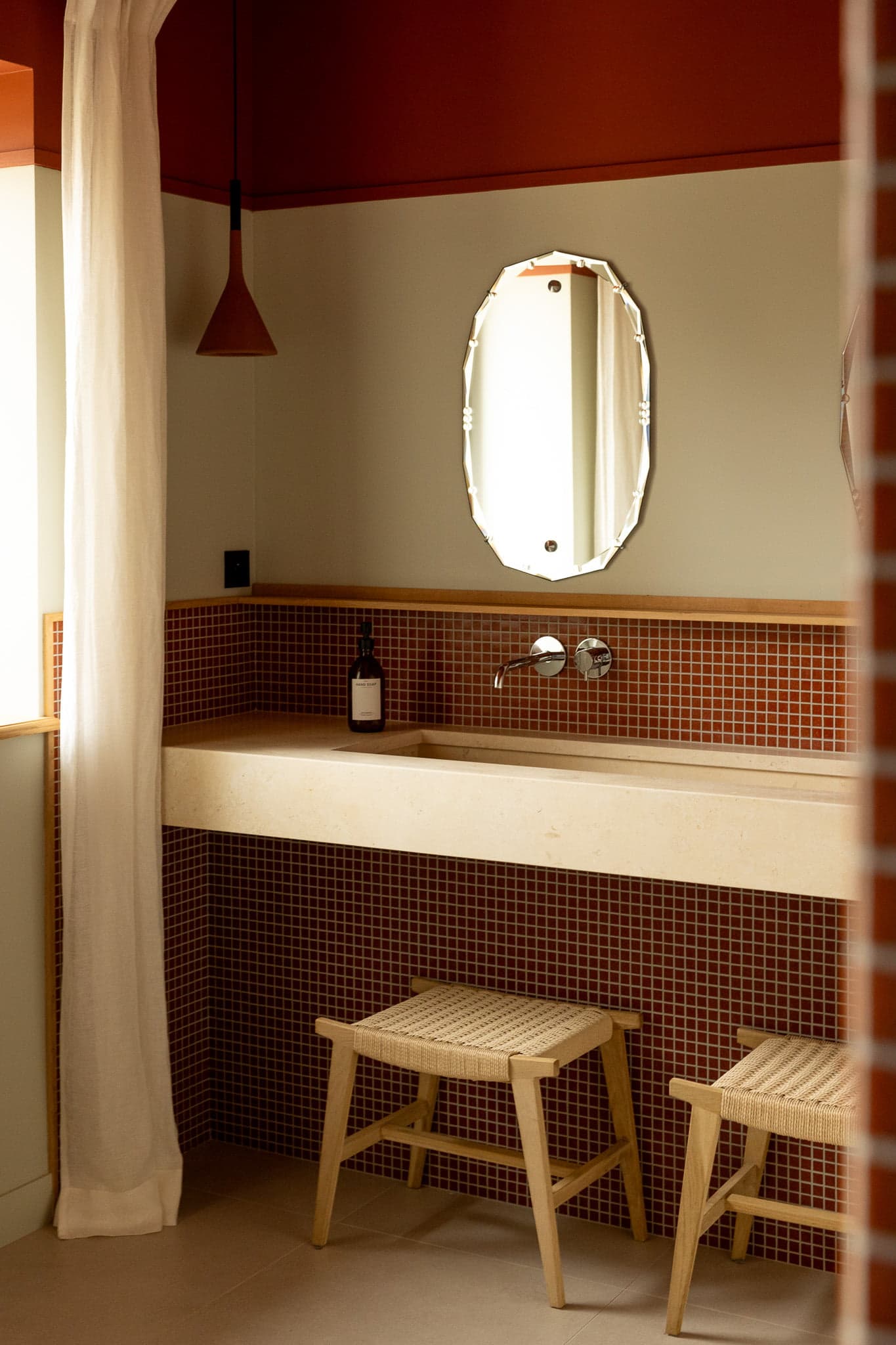 Une salle de bain confortable dotée d'un comptoir de couleur claire avec un lavabo intégré, des carreaux aux accents rouges, un miroir ovale monté et une bouteille de savon. Deux tabourets en bois se trouvent sous le comptoir et un rideau blanc est suspendu à une fenêtre voisine, adoucissant l'ambiance chaleureuse.