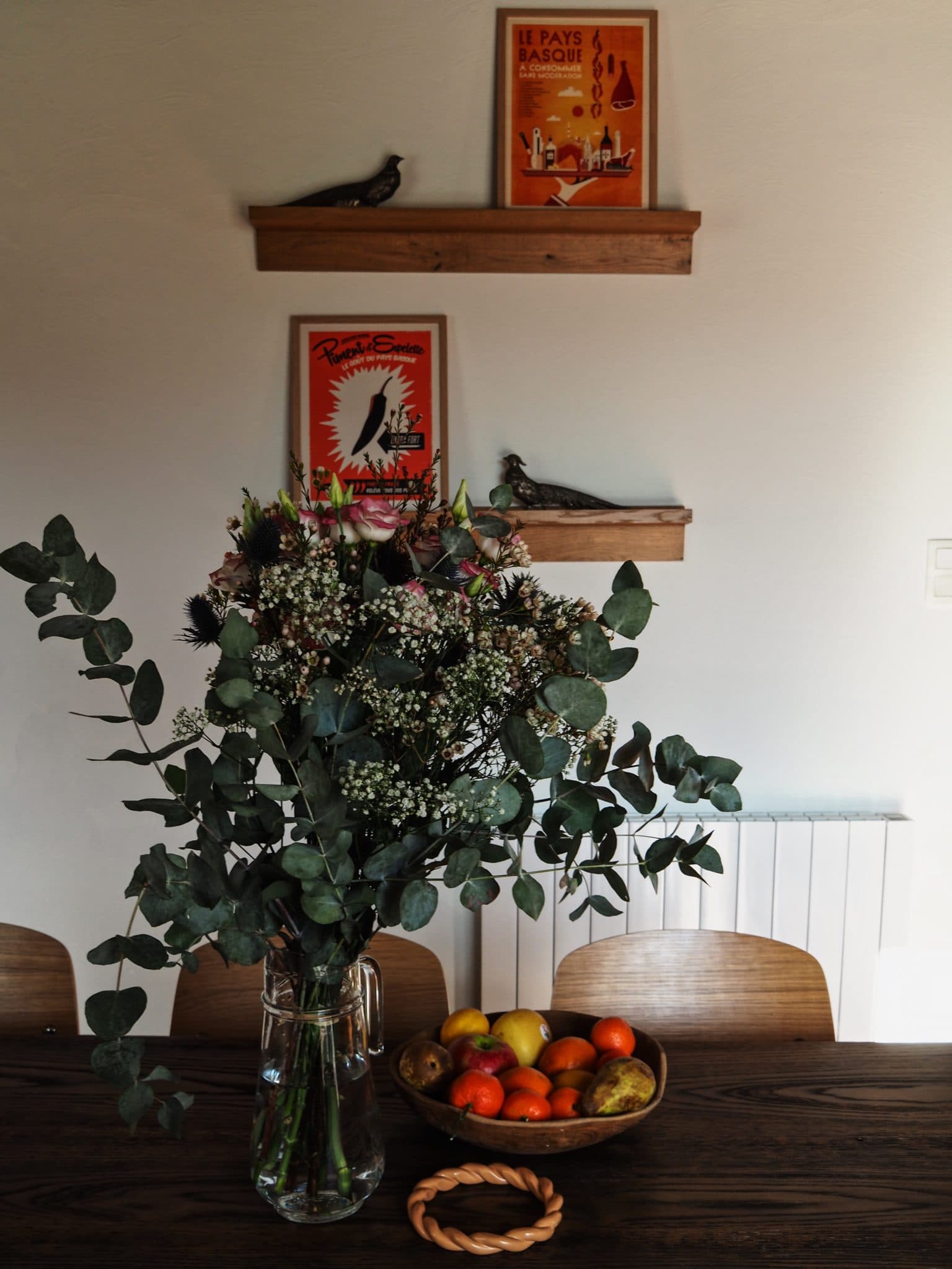 Un vase avec un grand bouquet de fleurs, dont de l'eucalyptus et du souffle de bébé, se trouve sur une table à manger en bois sombre. Un bol de fruits assortis se trouve à proximité. Deux petites étagères avec des figurines d'oiseaux et des affiches vintage sont montées sur le mur de couleur crème derrière.