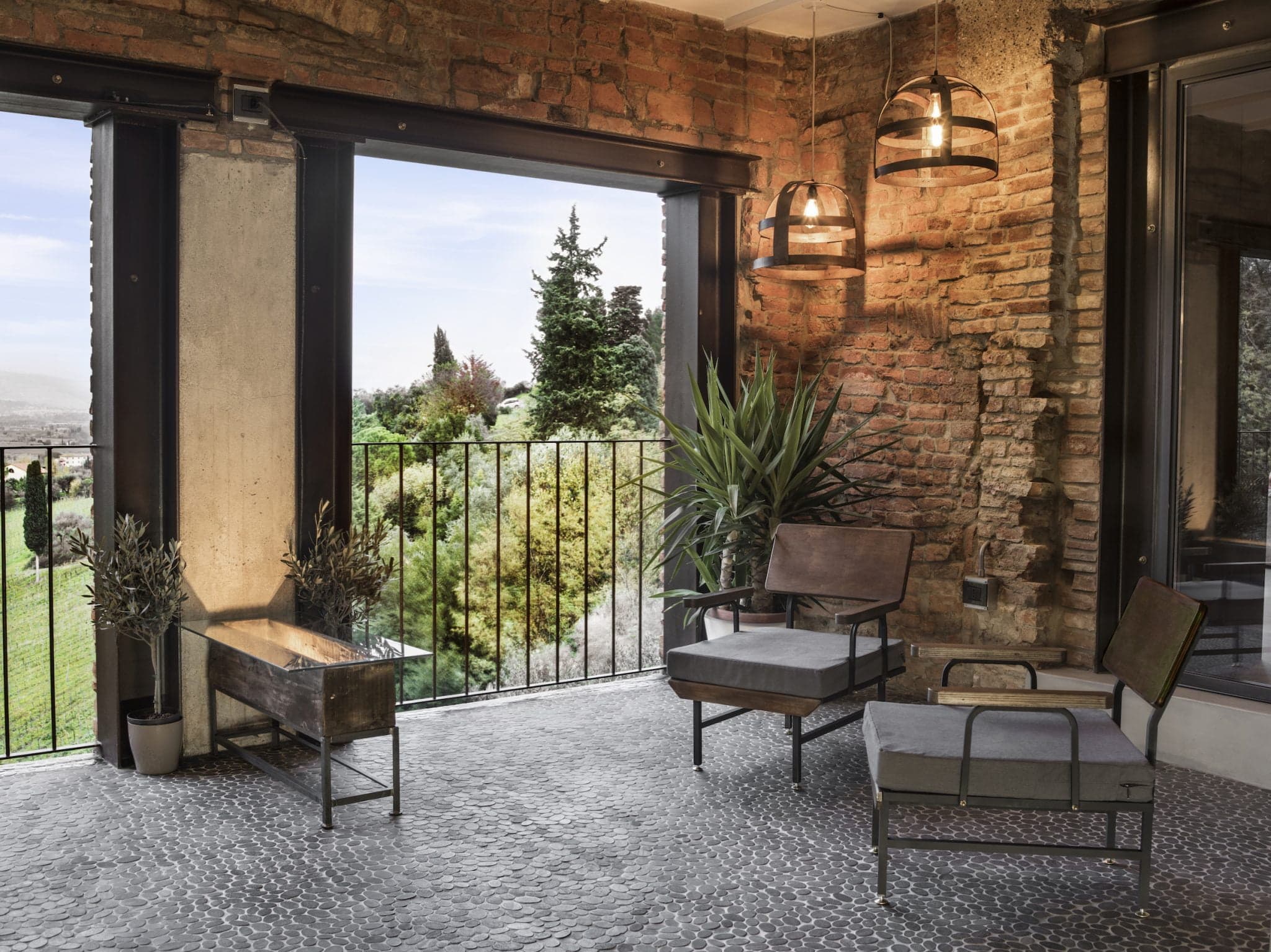 Un patio chaleureux est doté de deux chaises rembourrées et d'une petite table sur un sol en galets. Les murs en briques apparentes et les luminaires suspendus ajoutent un charme rustique. La verdure, à l'intérieur comme à l'extérieur, met en valeur l'espace. La vue donne sur un paysage luxuriant avec des arbres et une colline au loin.
